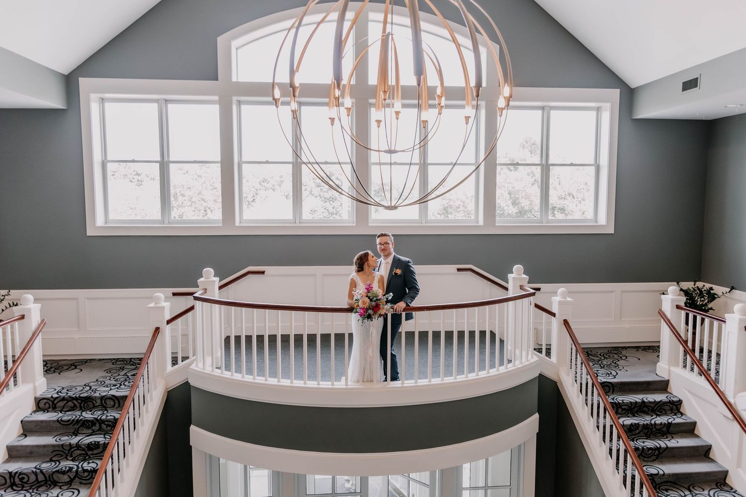 Wedding portrait of couple standing on elegant white balcony under large spherical chandelier.