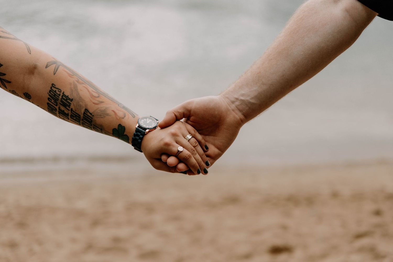 Close-up of two hands holding each other against a sandy beach background, showcasing tattoos and a wristwatch.