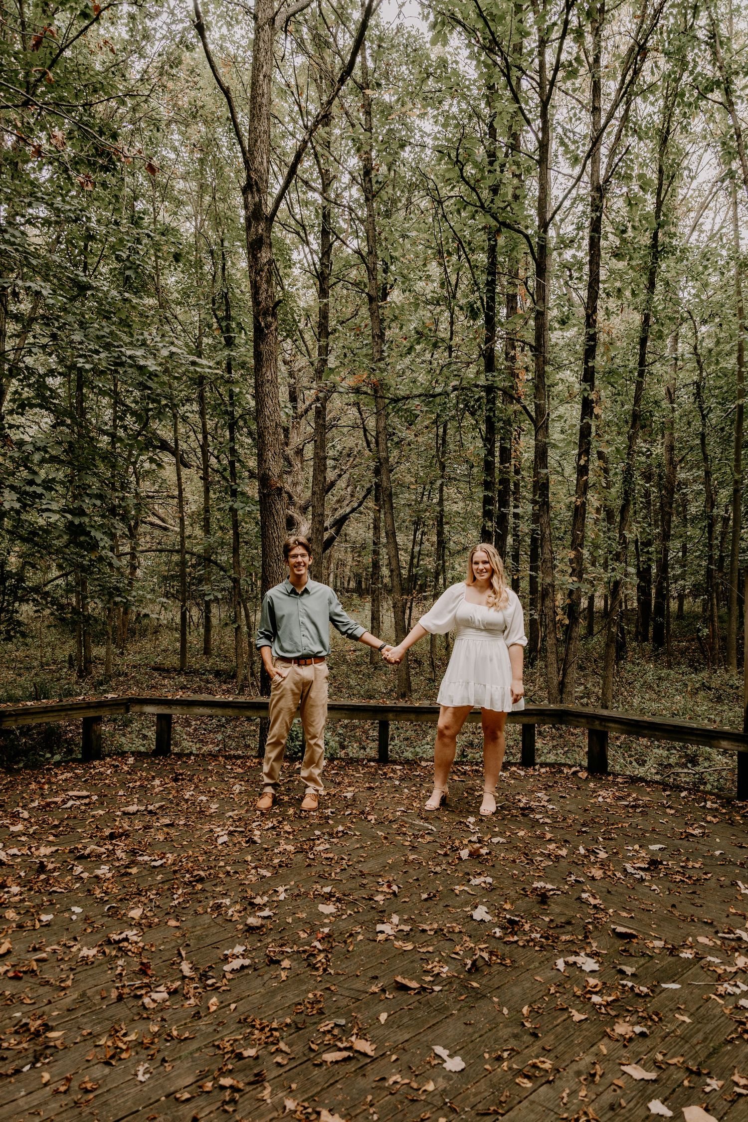 Couple holding hands while standing on wooden platform in forest setting.
