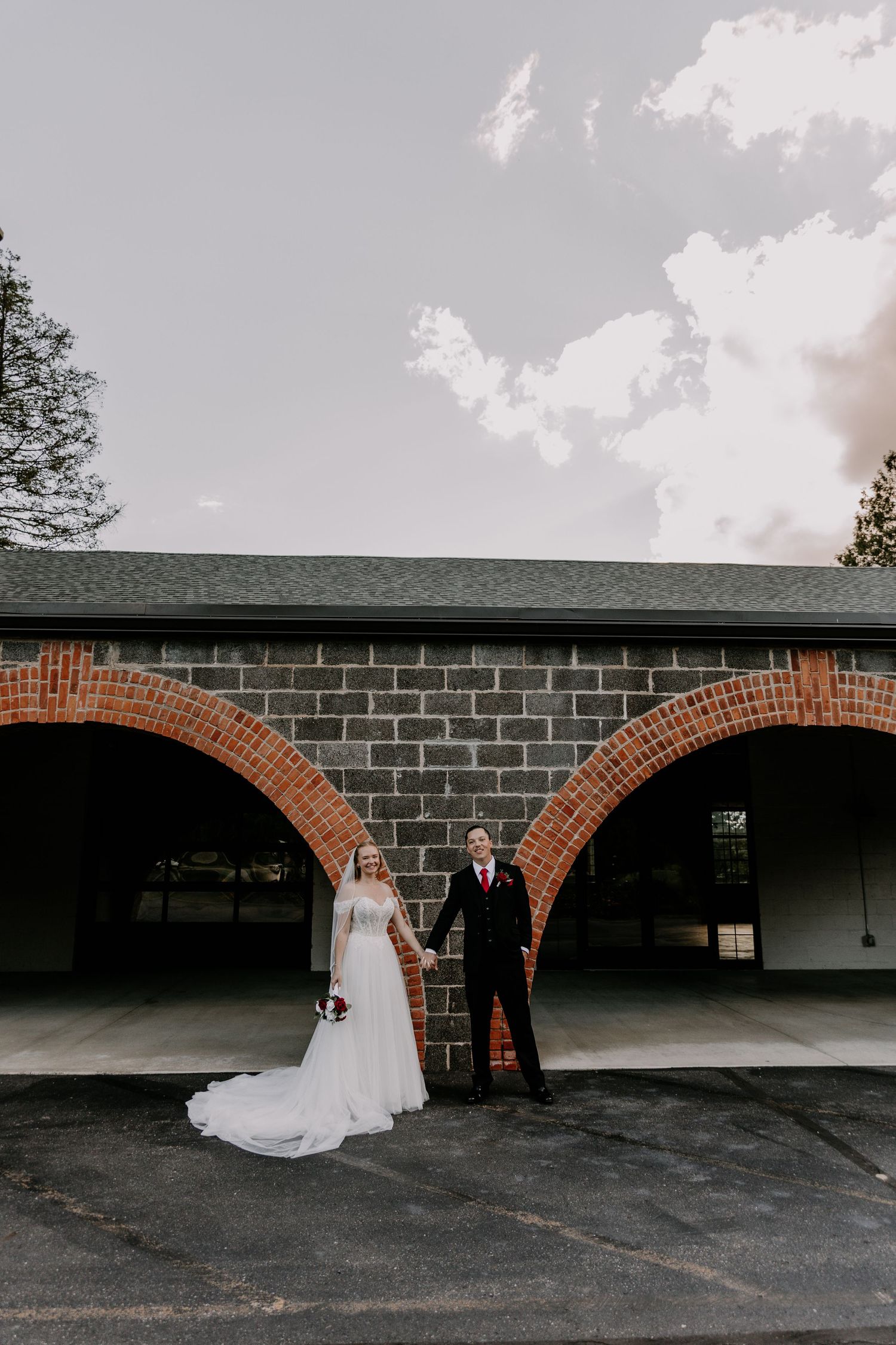 Bride and groom hold hands between brick arches at their wedding venue, bride in flowing white gown with bouquet.