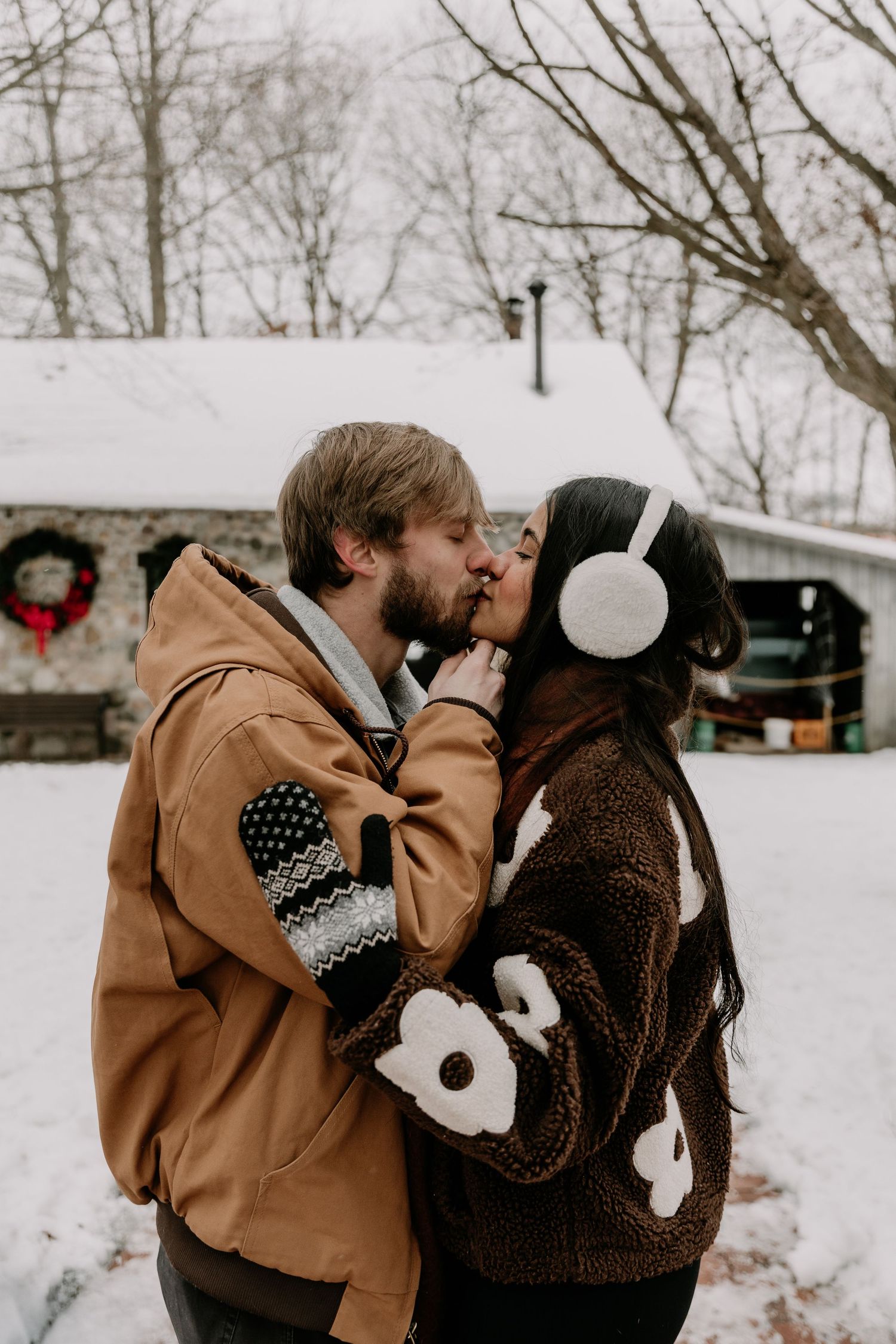 A couple plays together in the snow during a romantic winter photoshoot with a rustic backdrop.