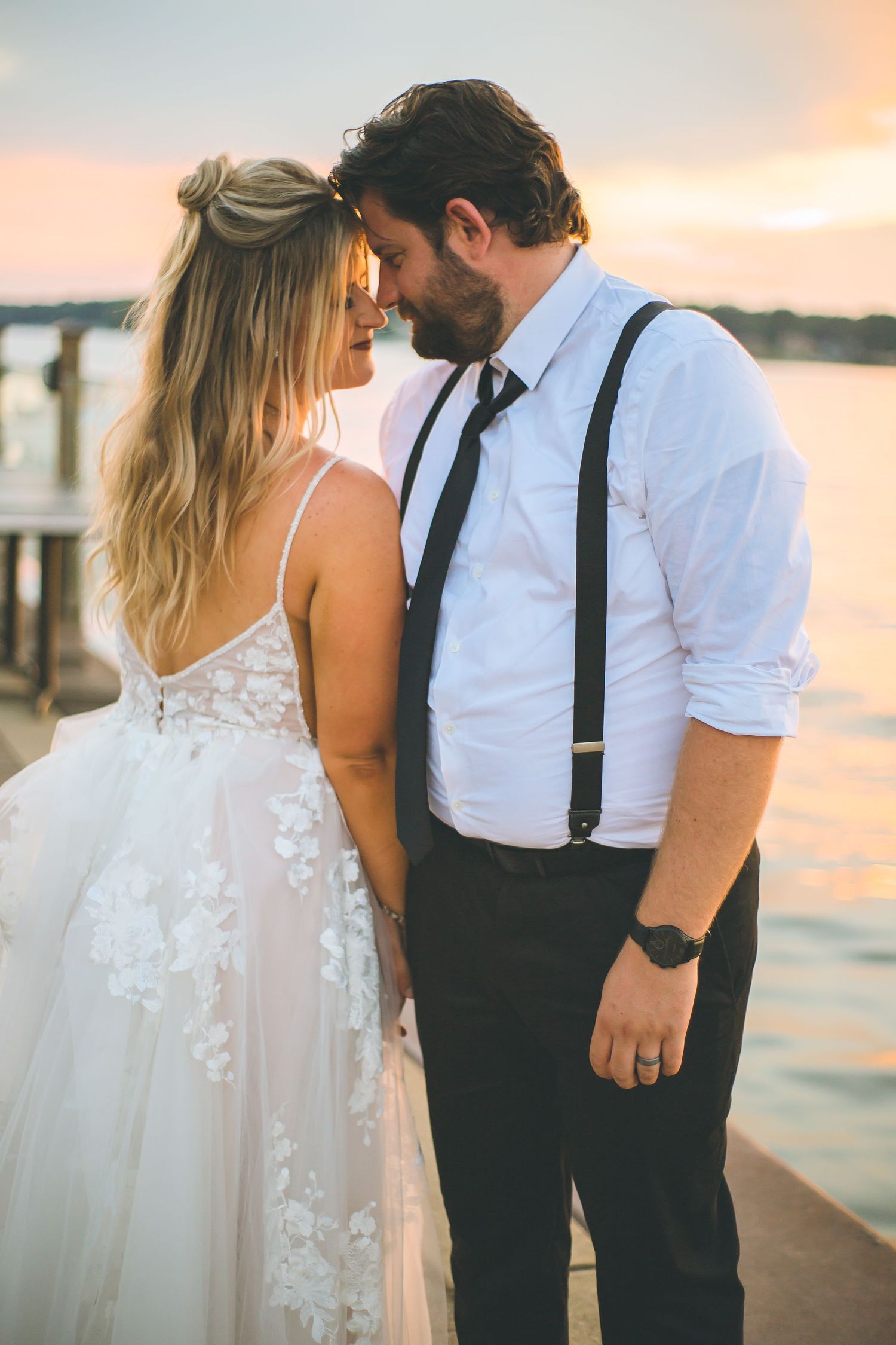A wedding couple in white and suspenders poses against a golden sunset by the water.