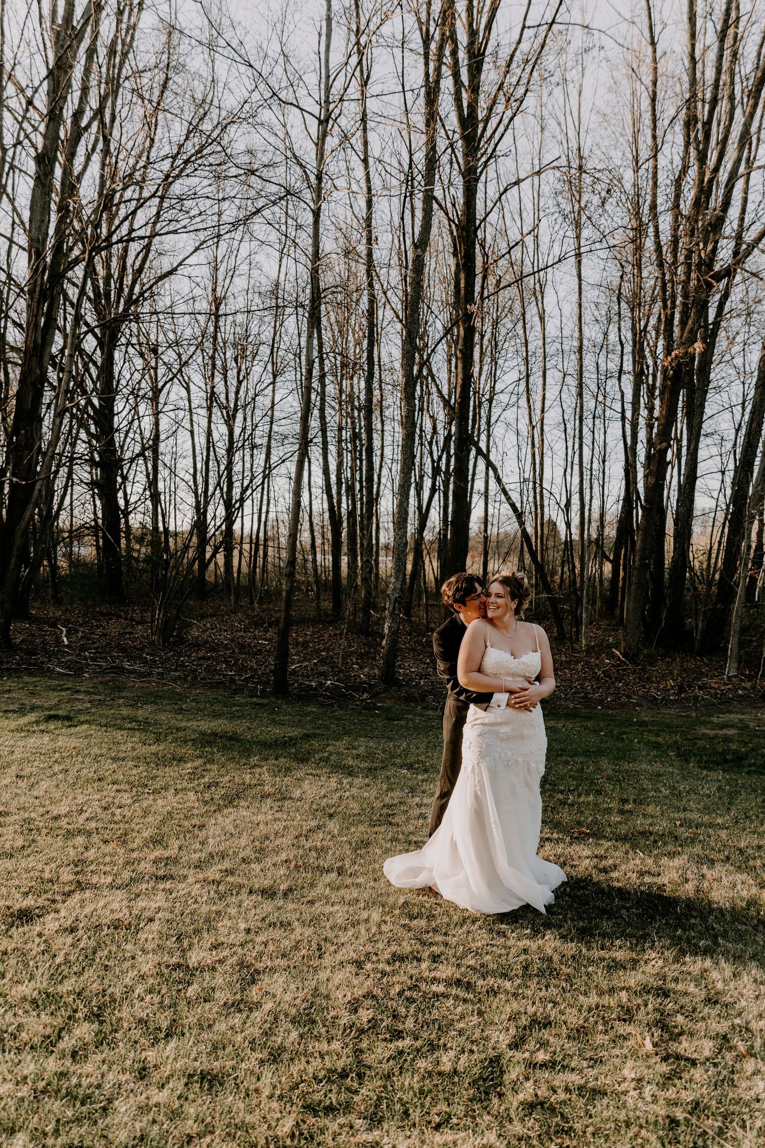 Couple in wedding attire embracing in field with bare winter trees in background.