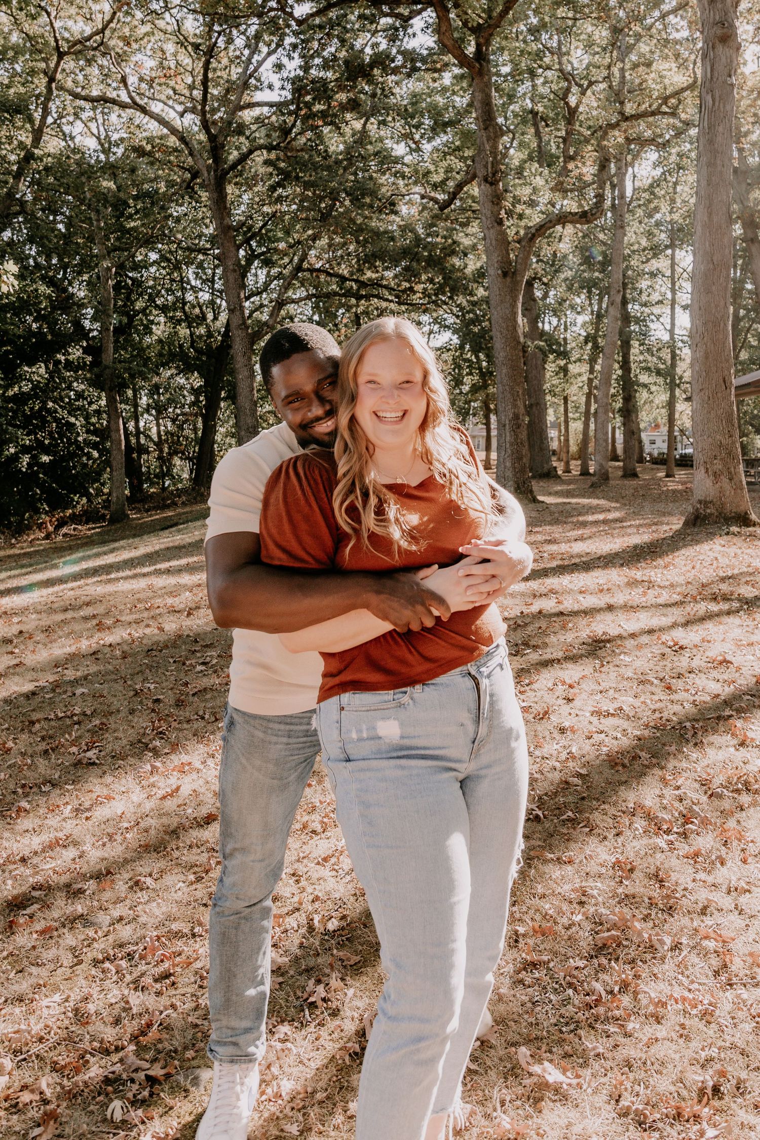 A couple embraces joyfully in a sunlit wooded area with fallen autumn leaves on the ground.