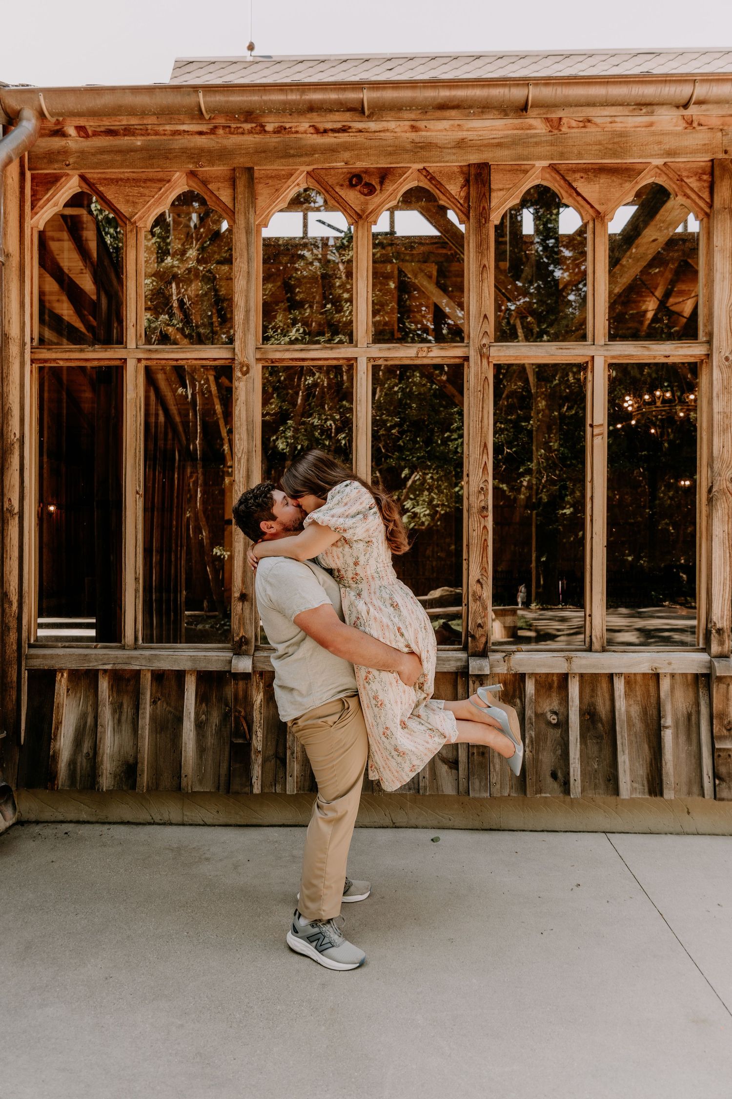 Man lifting and kissing woman in front of a rustic wooden building with arched windows outdoors.