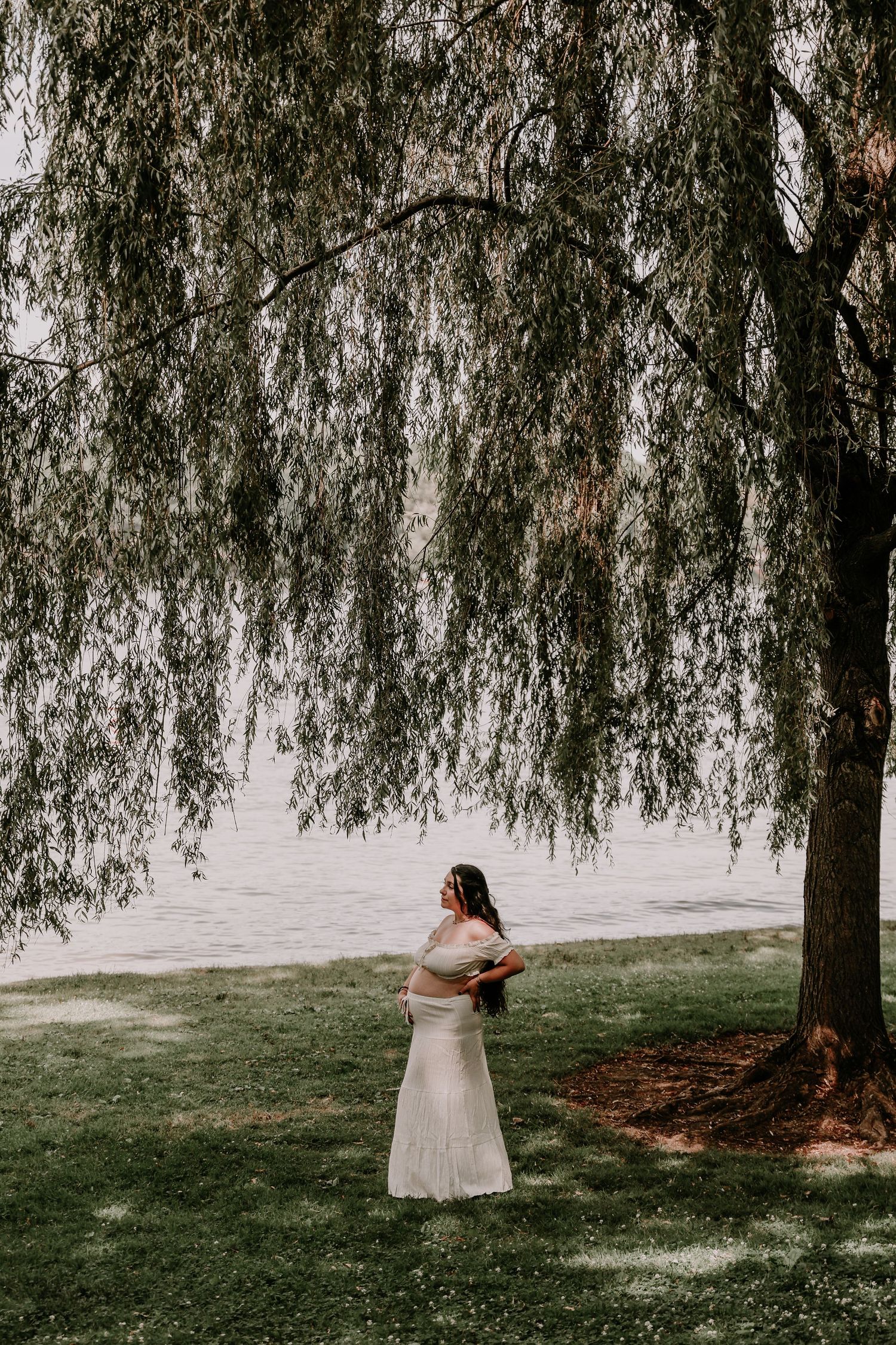 Pregnant woman in white dress standing under weeping willow tree near waterfront.