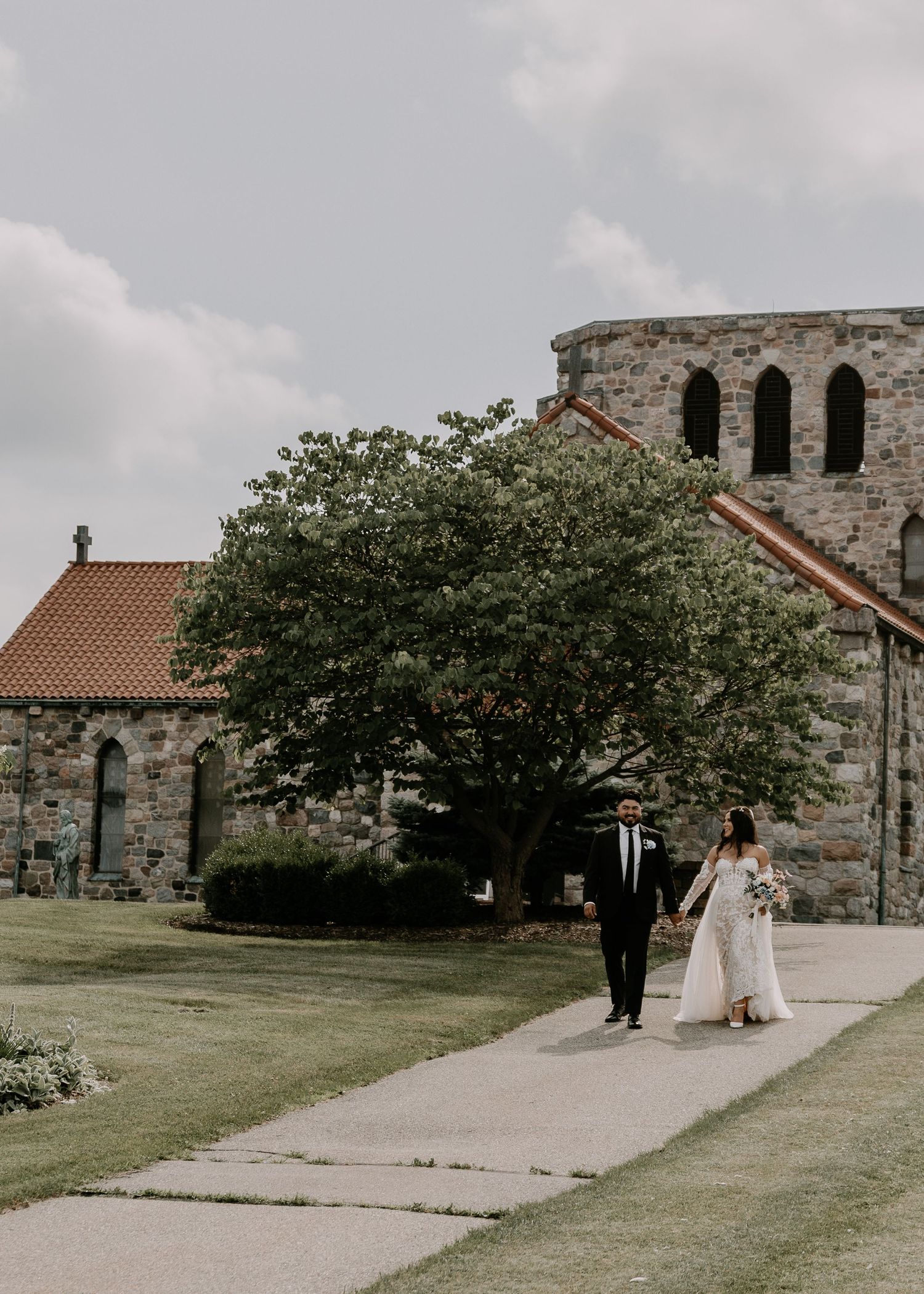 Bride and groom walk along stone path outside historic church with arched windows and blooming tree on cloudy day.