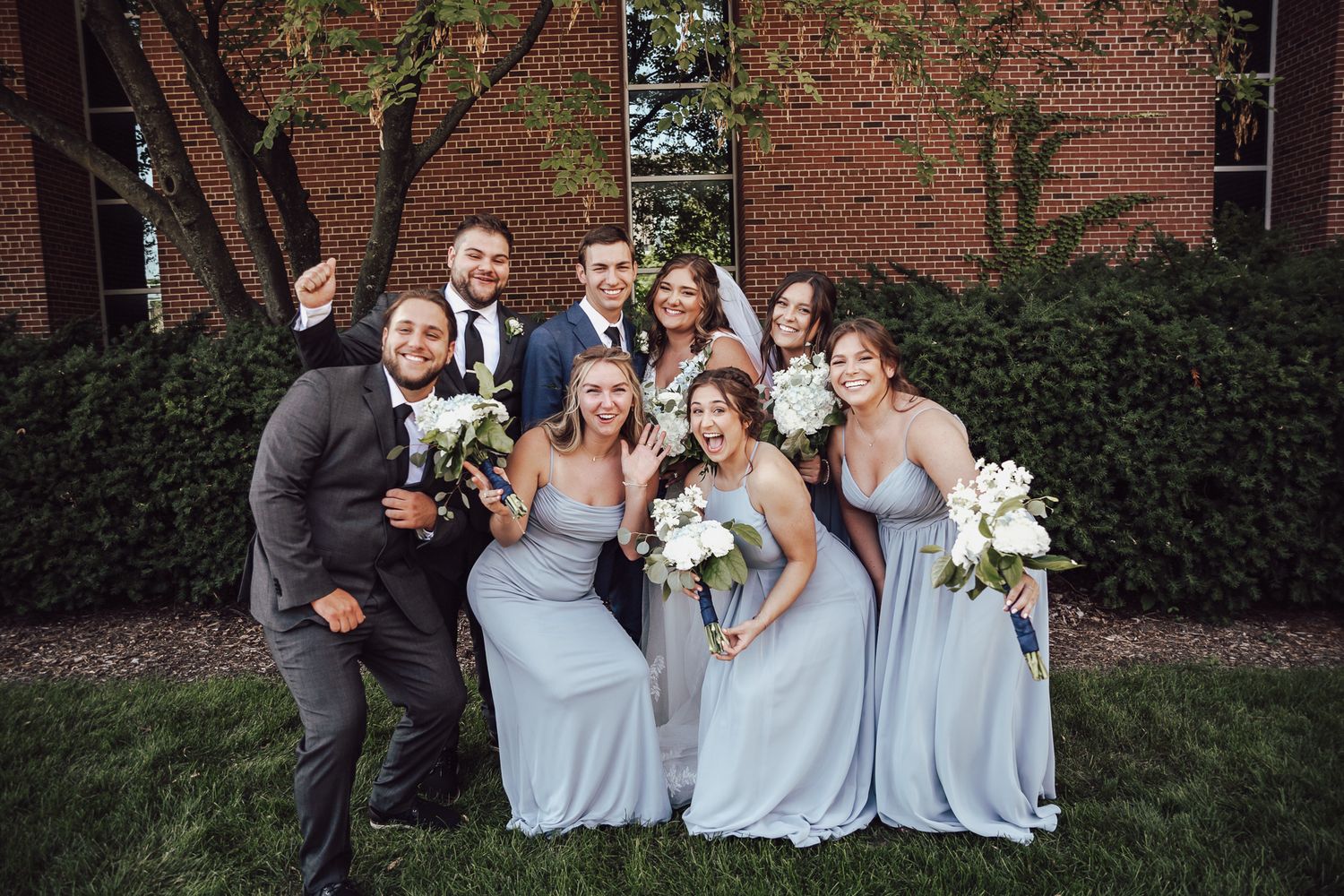 Wedding party poses together for a fun group photo in front of a brick building.