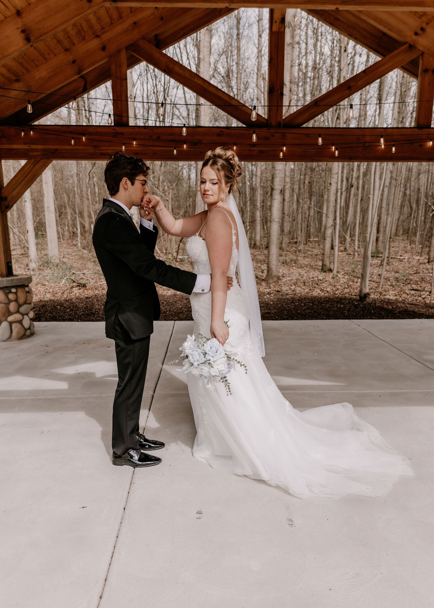 Wedding couple dancing together under a rustic wooden pavilion surrounded by bare trees.