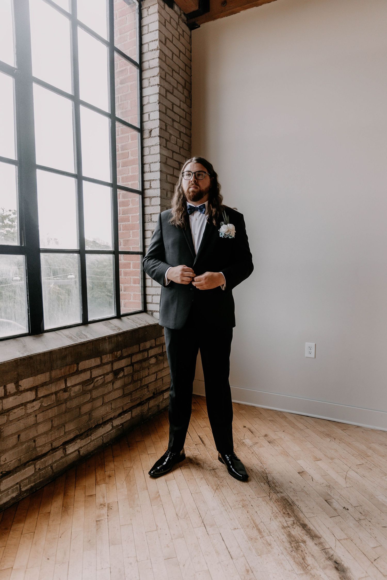 A formal portrait of someone in a bowtie and tuxedo standing next to floor-to-ceiling windows.