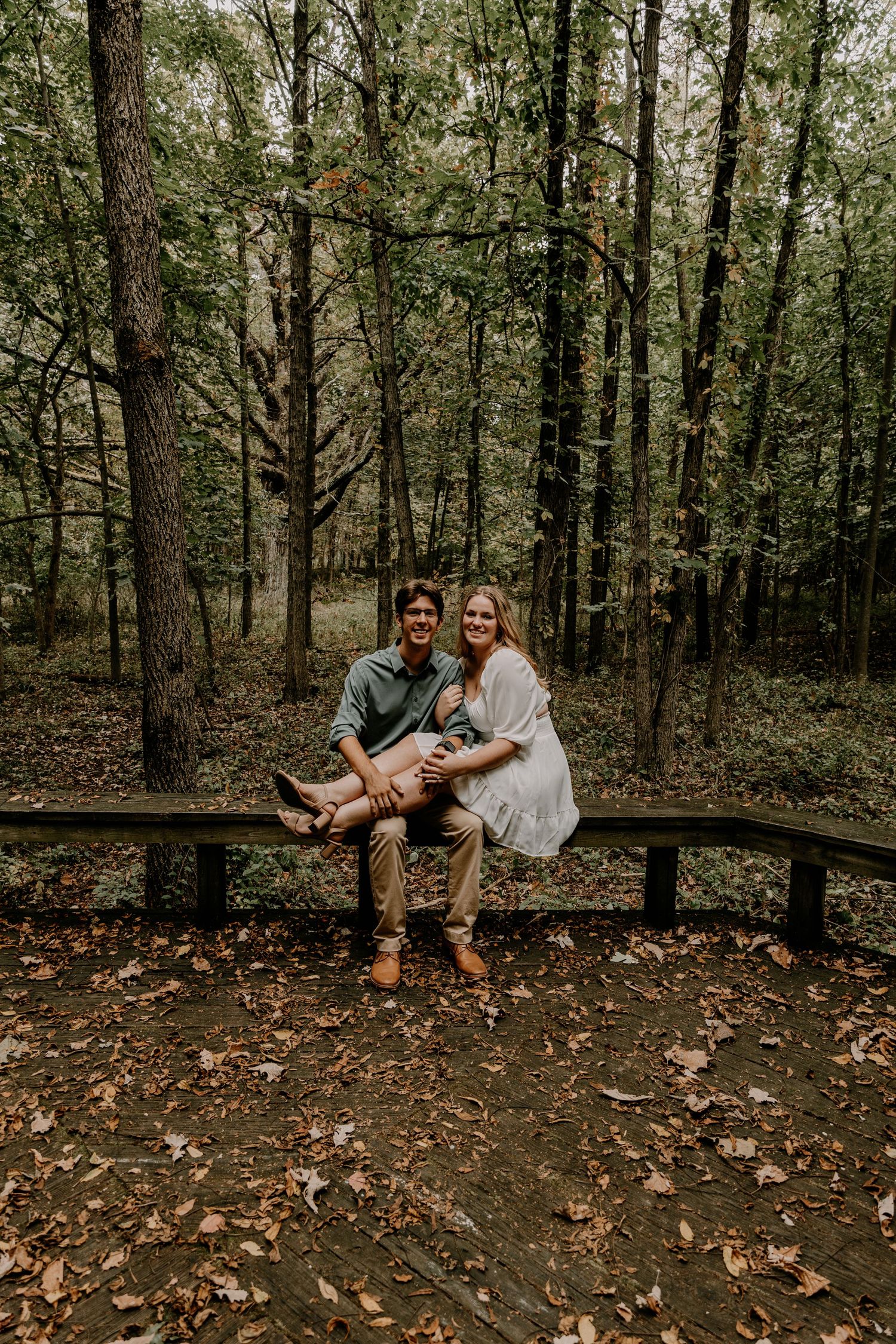 A couple sits intimately on a wooden bench surrounded by a forest of tall trees on an autumn day.