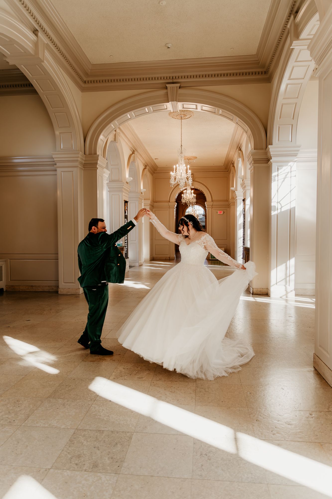 A couple dancing in an elegant hallway with sunlight streaming through arches and crystal chandeliers above.