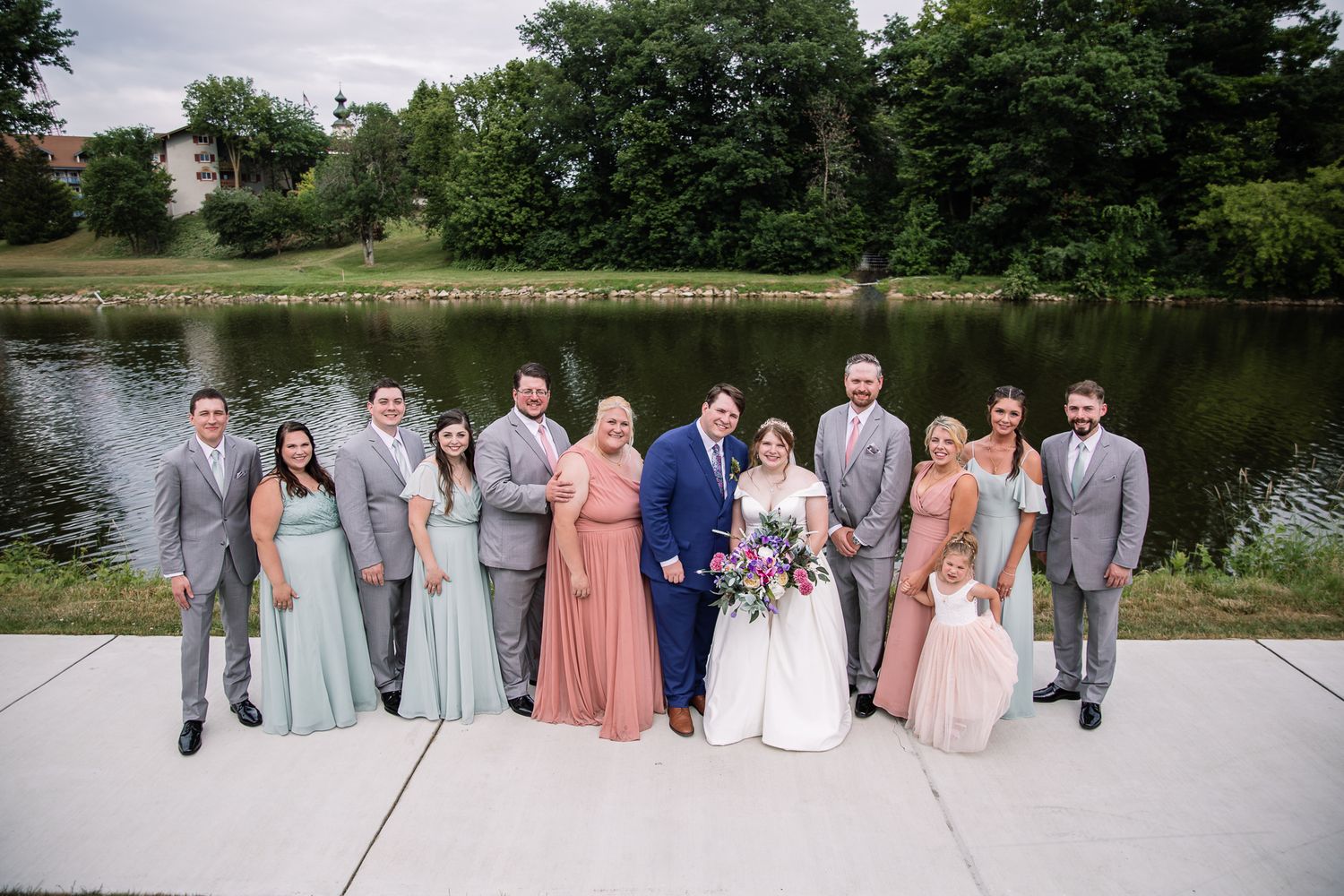 Wedding party poses by a lake wearing coordinated pastel and gray attire.