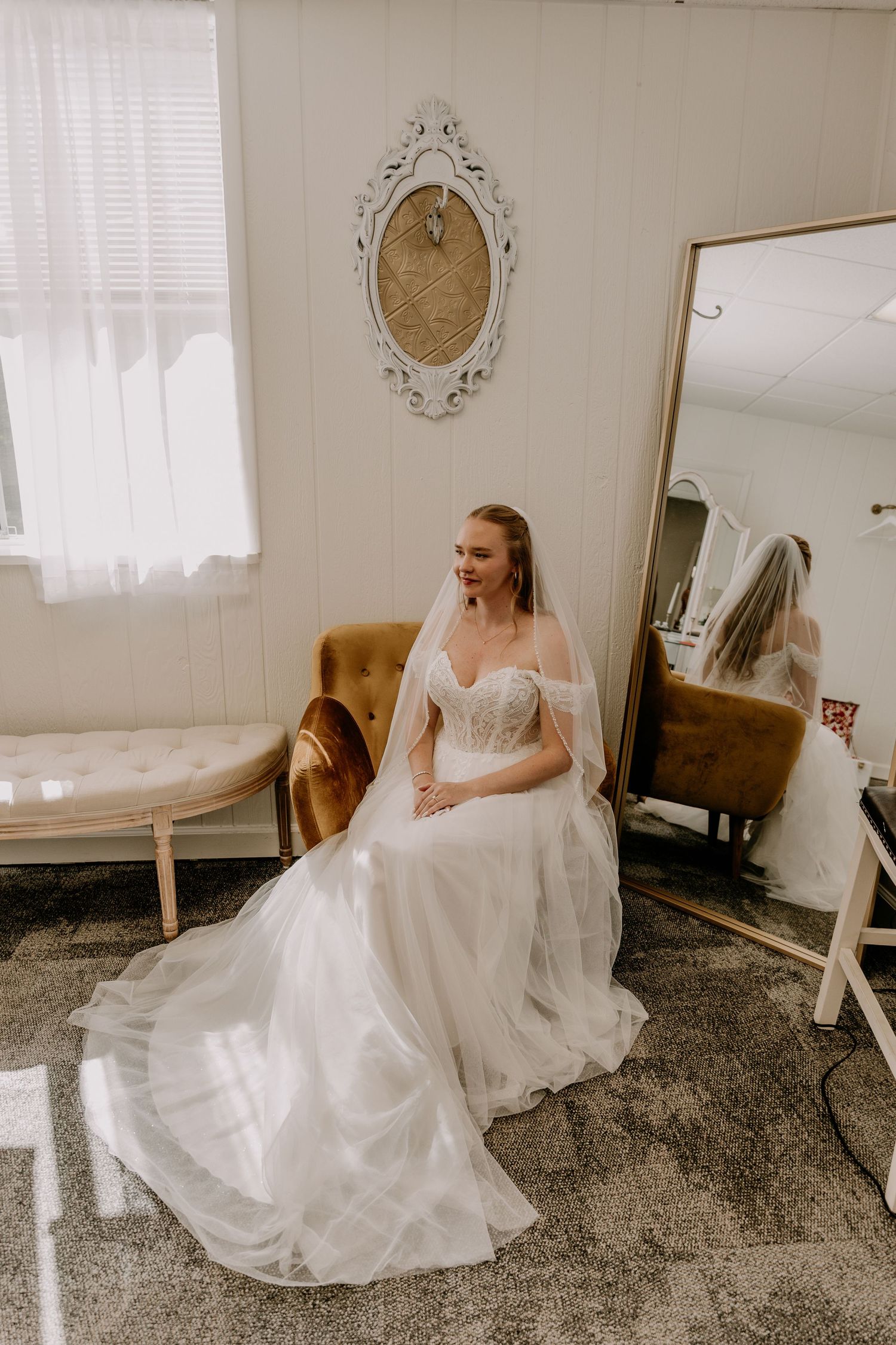 Bride in white off-shoulder wedding gown with veil sits in bridal suite near mirror, smiling softly.