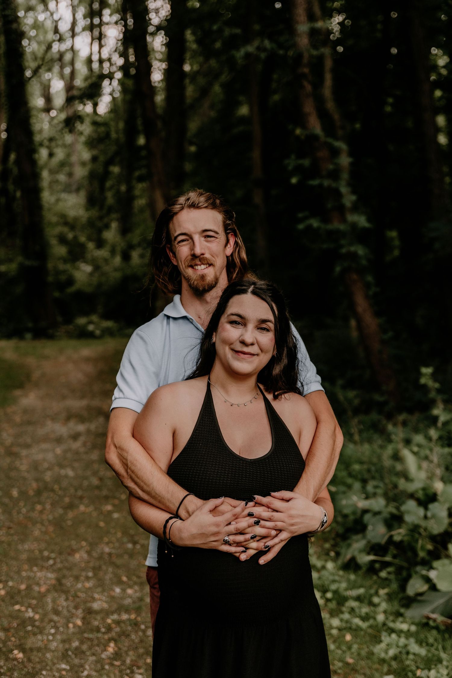 A couple embraces on a forest path, woman in black dress, man in blue shirt, smiling warmly among lush green trees.