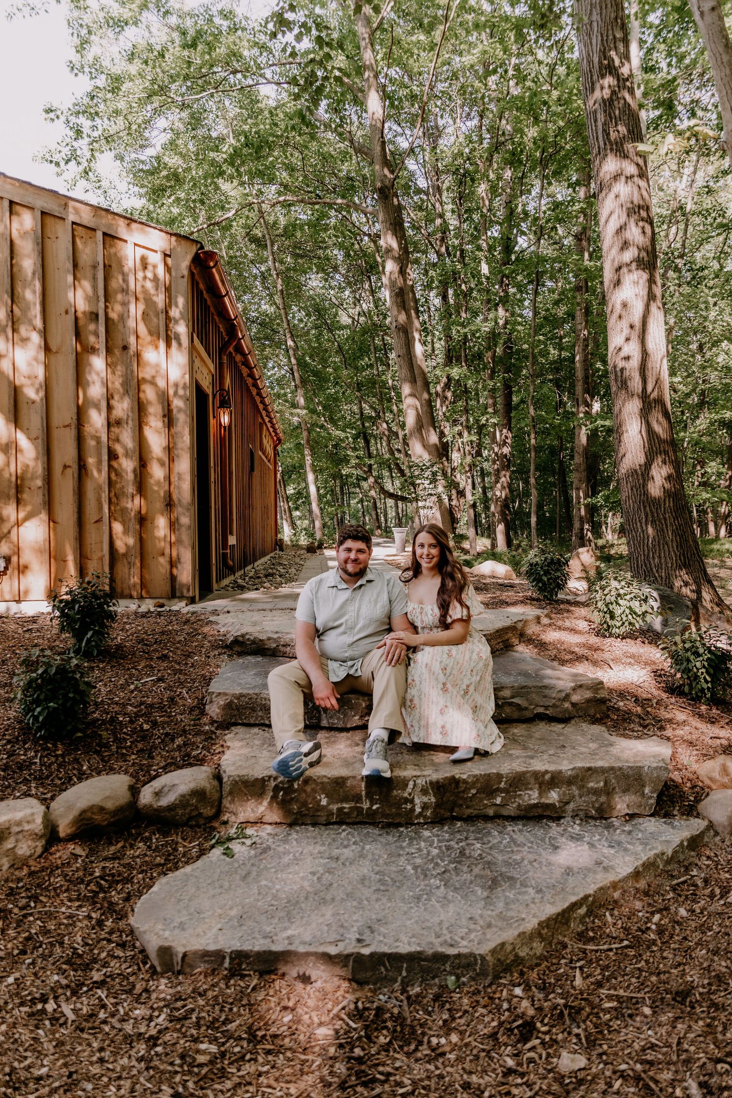 Couple sitting on stone steps beside a rustic wooden barn surrounded by tall trees in a wooded setting.