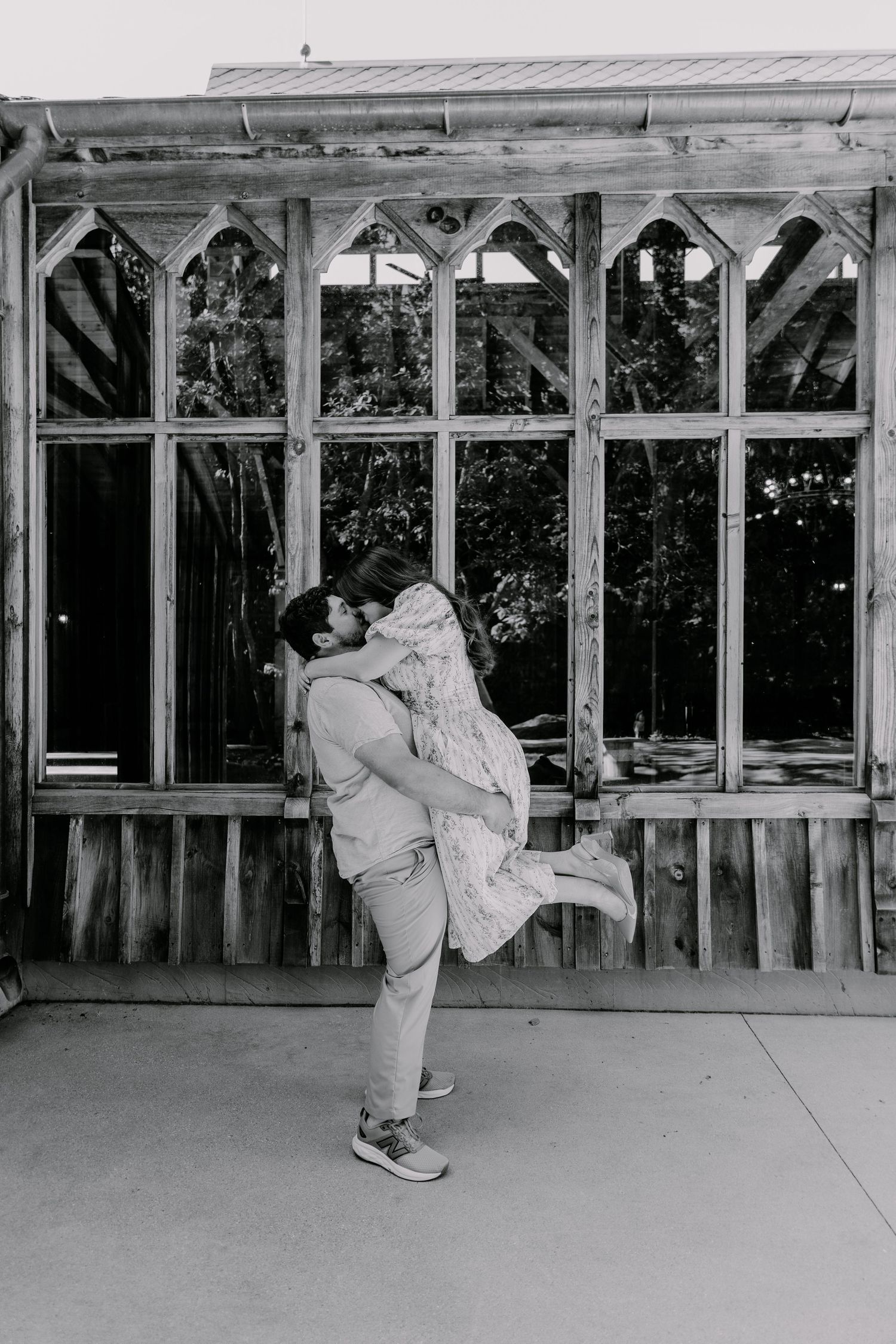 Black and white photo of a man lifting his partner in a romantic embrace outside a rustic wooden greenhouse.