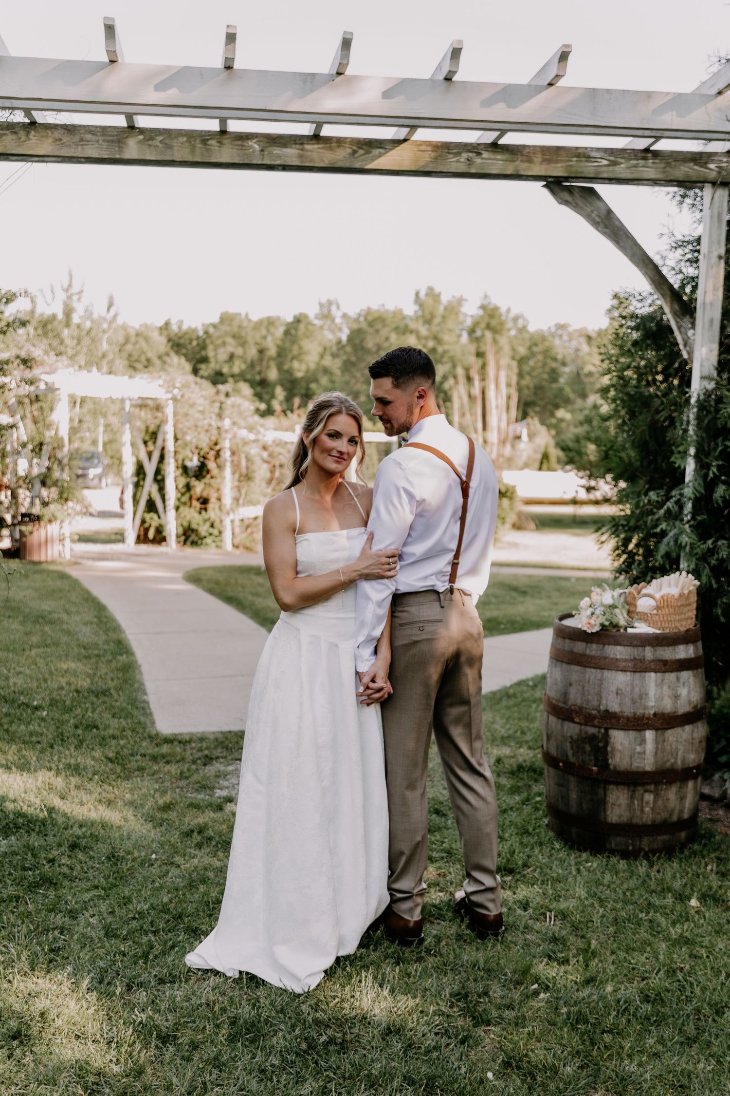 Bride in white gown and groom in suspenders share a romantic moment under a rustic pergola at an outdoor wedding.