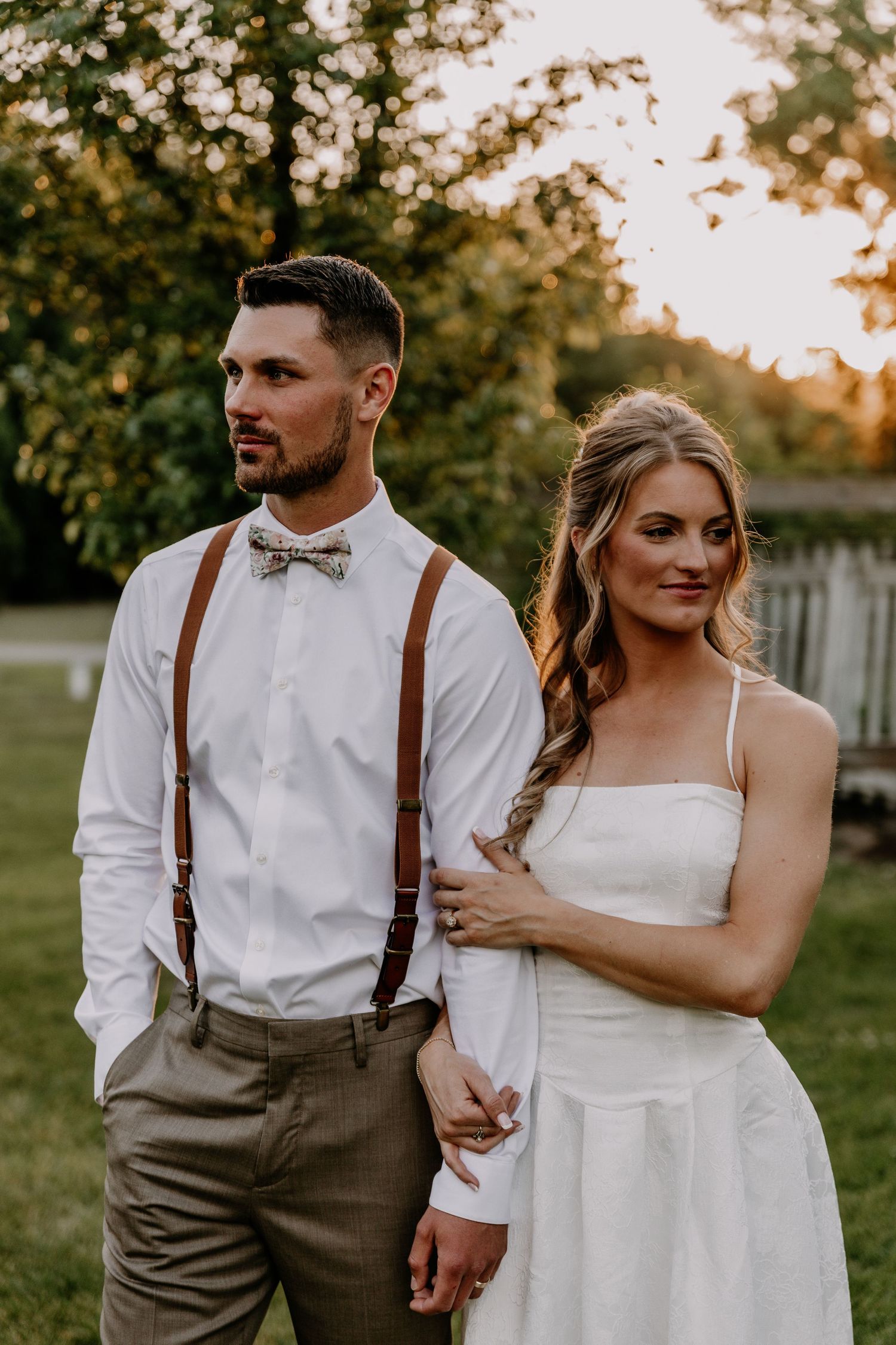 Wedding couple stands together outdoors at sunset, groom wearing brown suspenders and floral bow tie with bride.