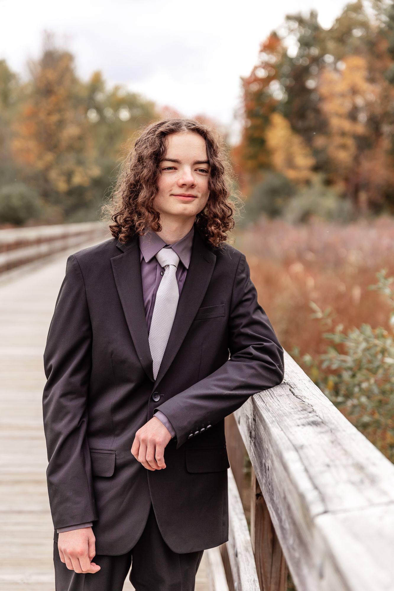 Portrait of person in dark suit leaning against railing on wooden bridge in fall scenery.