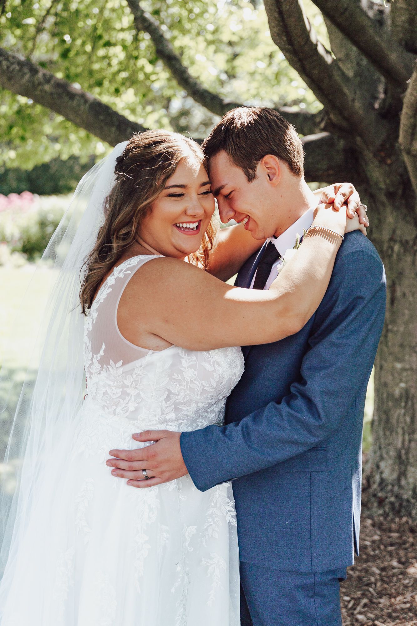 Bride and groom share tender moment while embracing outdoors under flowering tree.