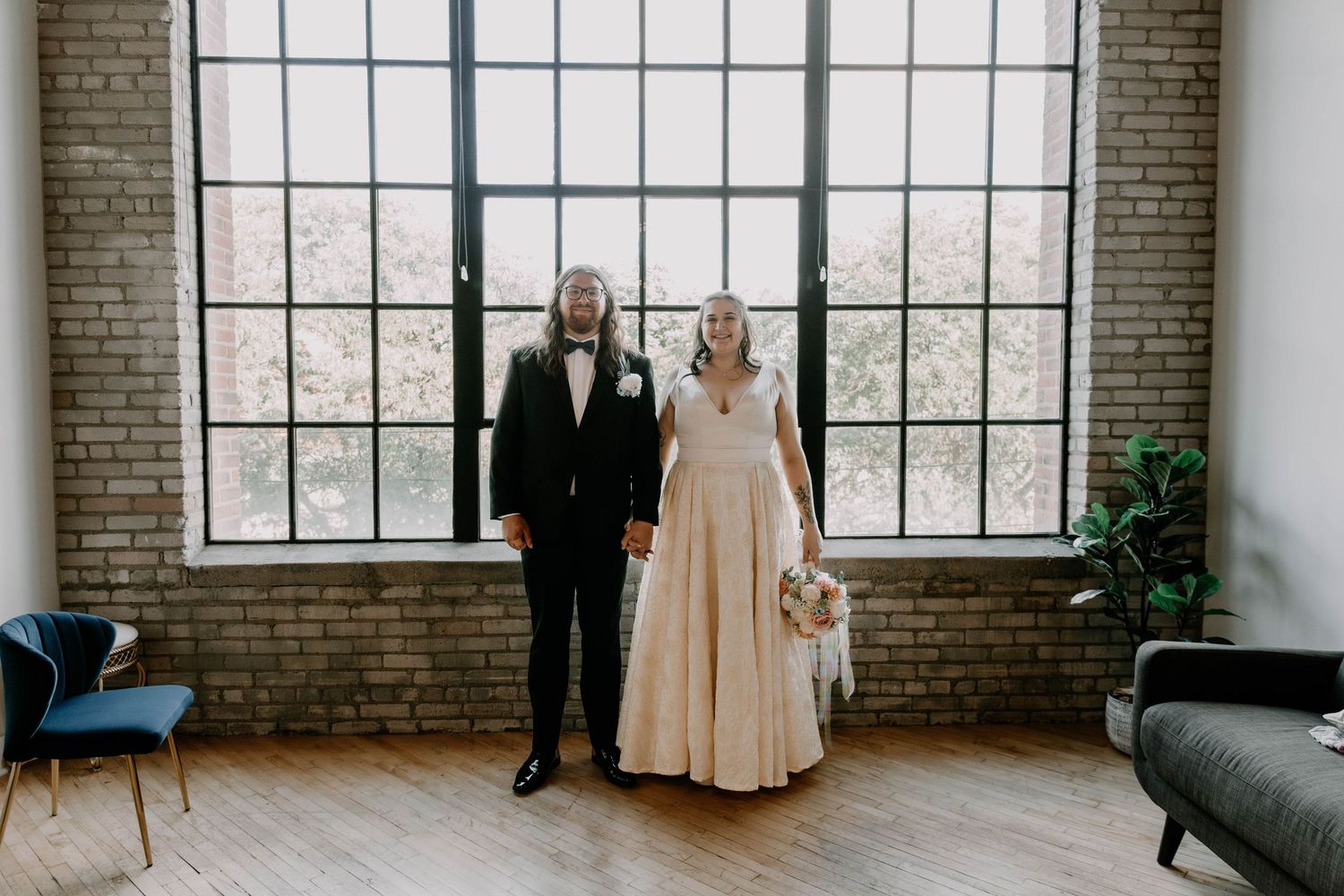 A couple poses formally in wedding attire against bright industrial windows and brick walls.