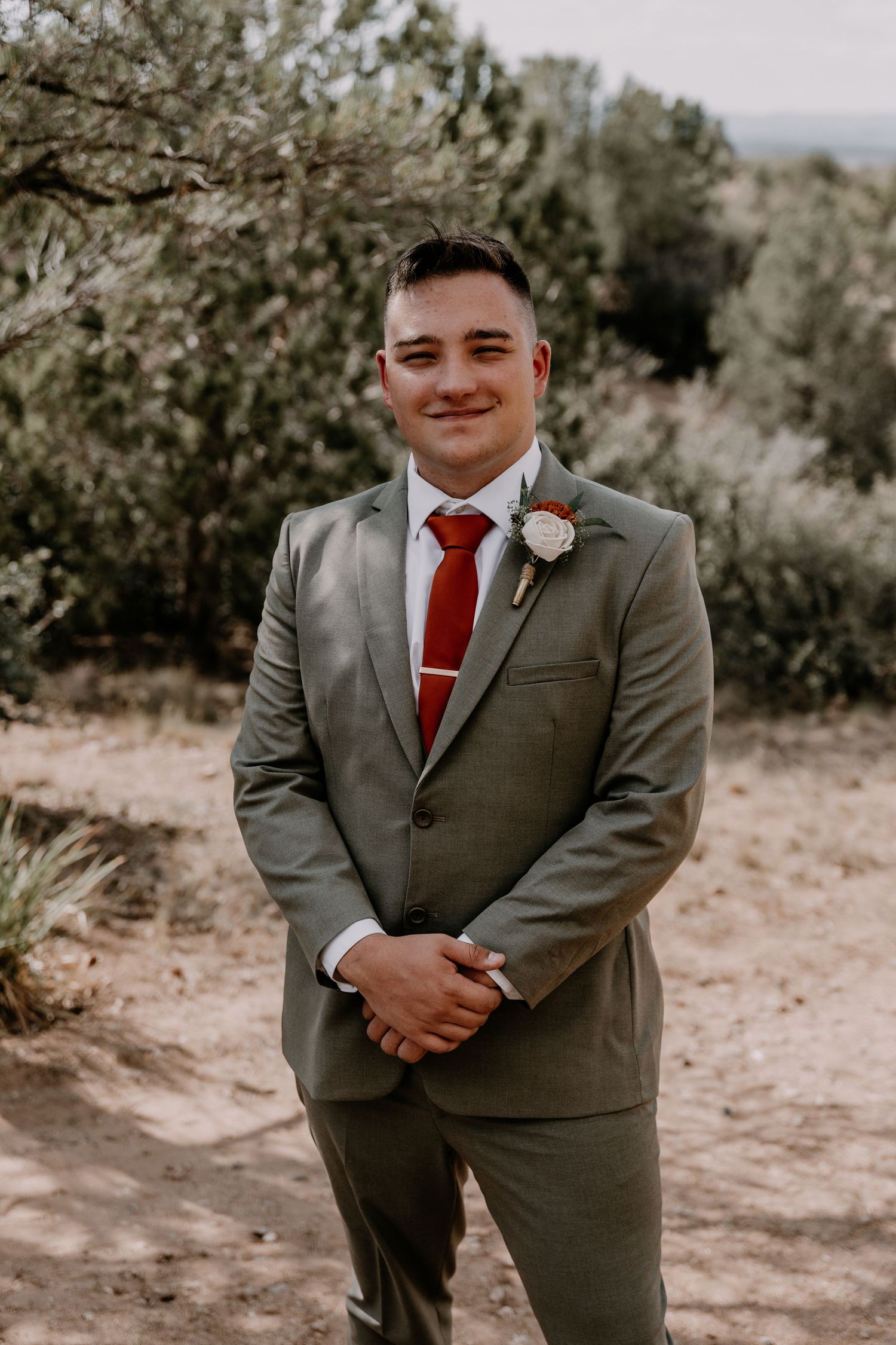 A groomsman in a grey suit with red tie poses in a desert setting.