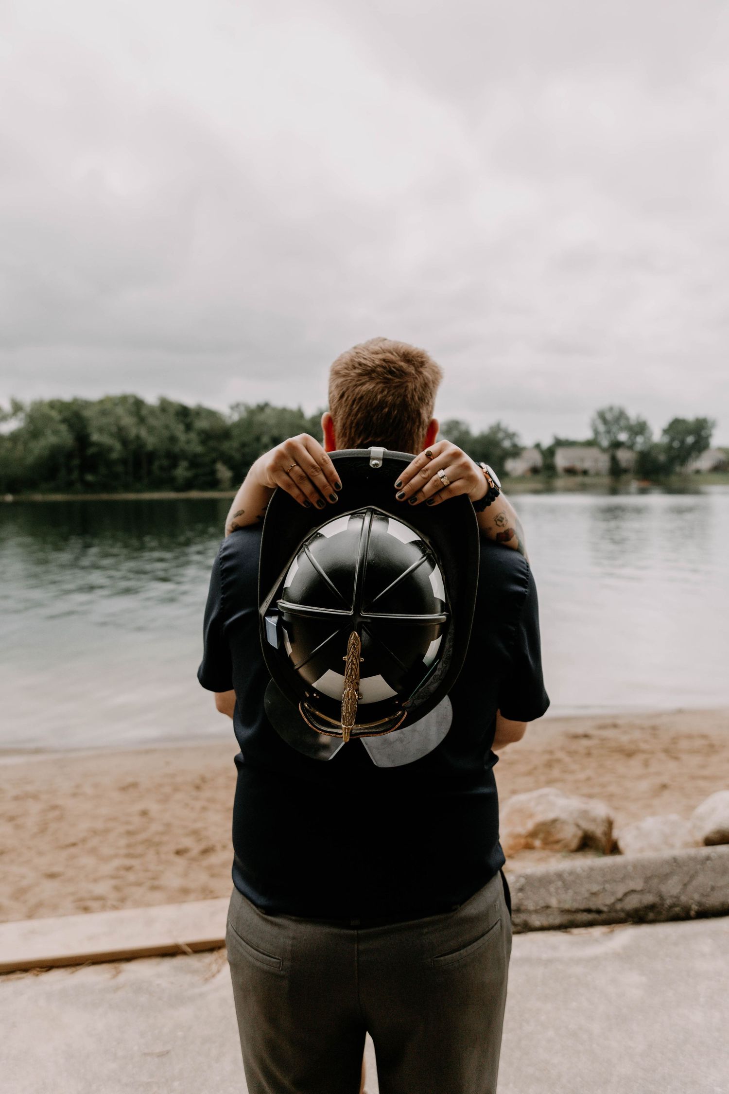 Person wearing black shirt holds backpack while facing lake on cloudy day.