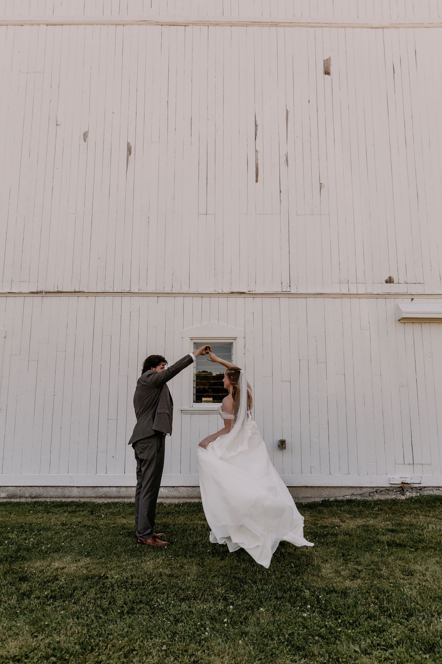 Groom twirls bride in white gown outside a rustic white barn wedding venue.
