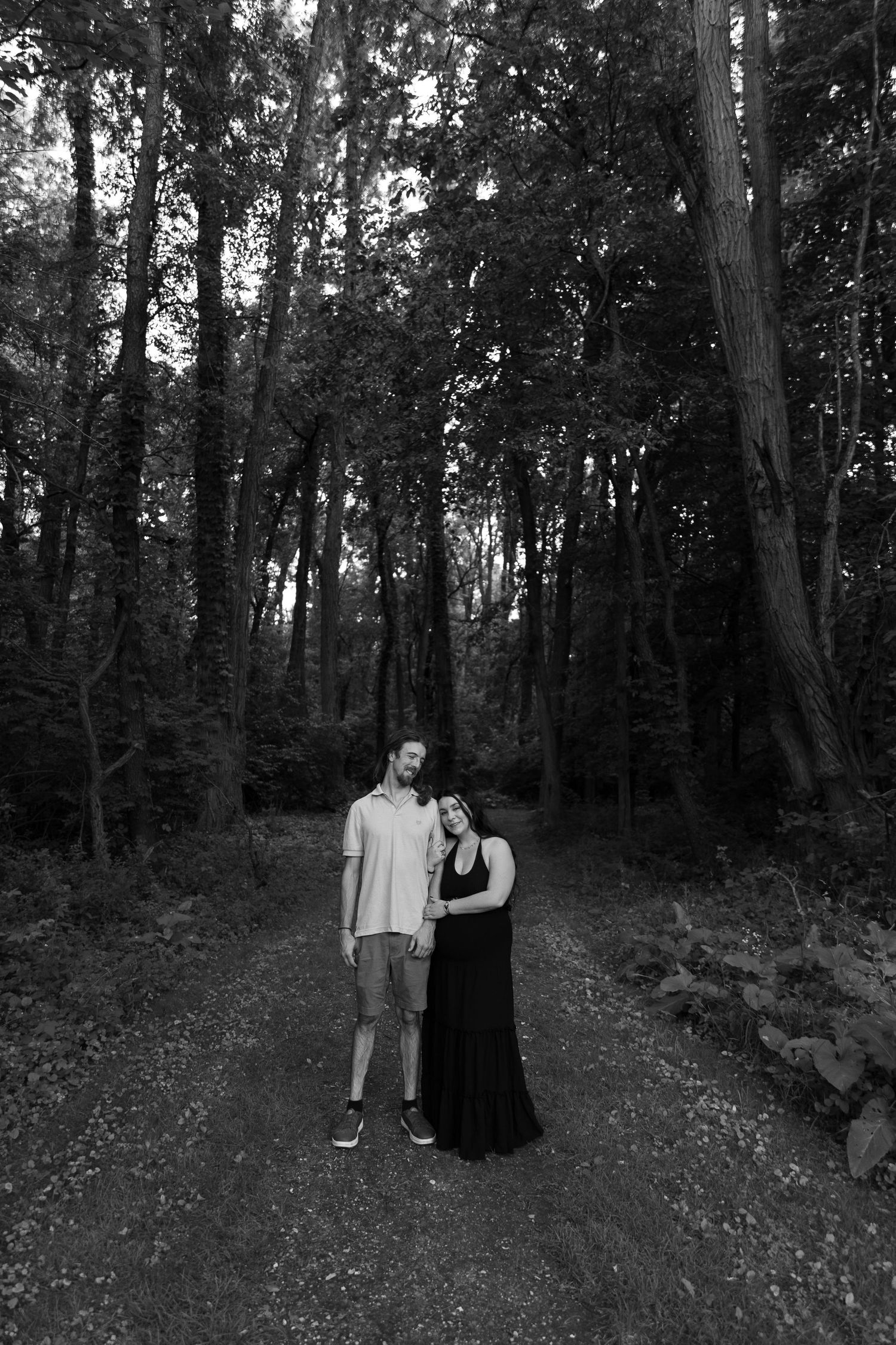 Couple embracing on a forest path surrounded by tall trees in a romantic black and white photo.
