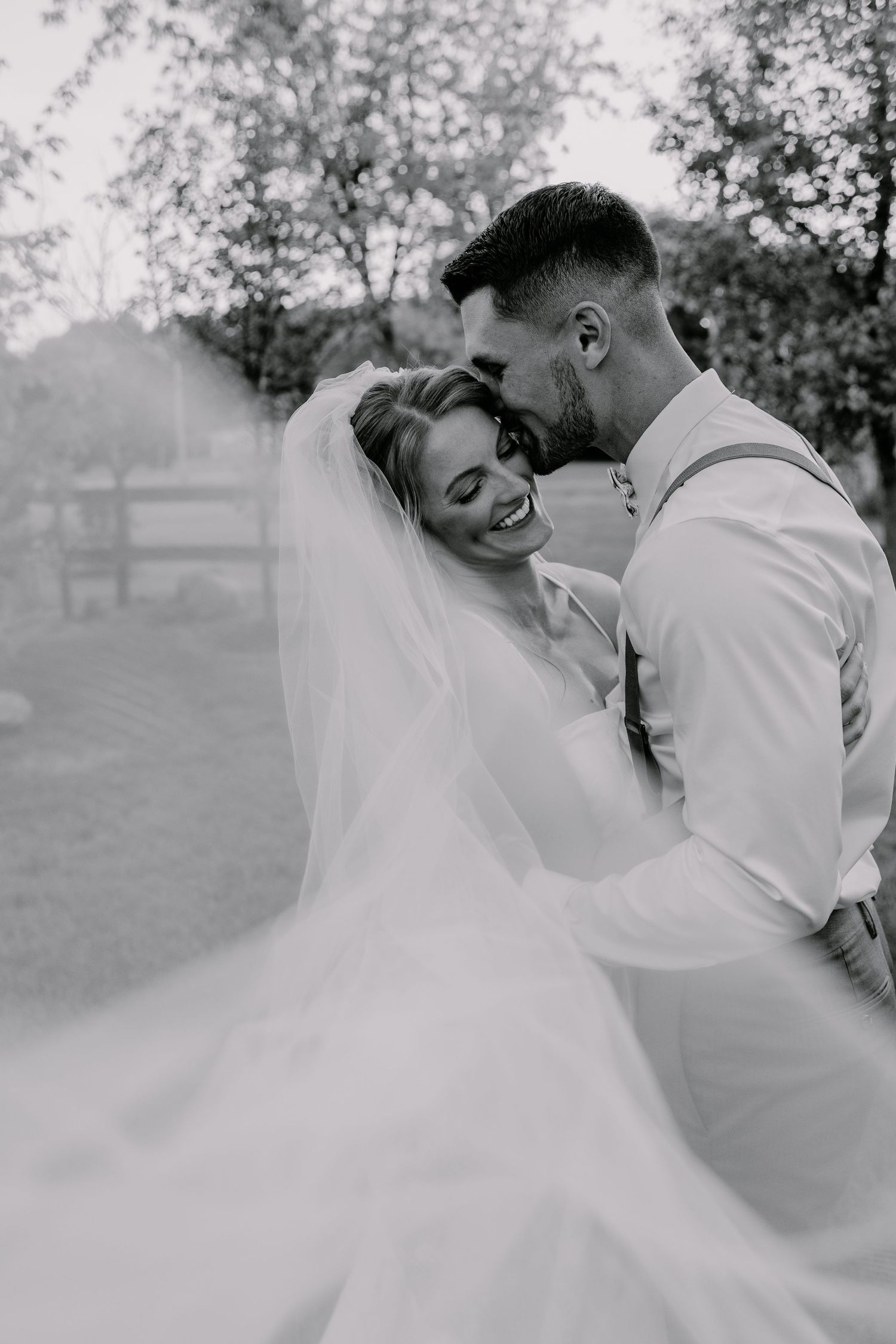Black and white wedding photo of smiling bride in veil and groom sharing a tender moment outdoors.