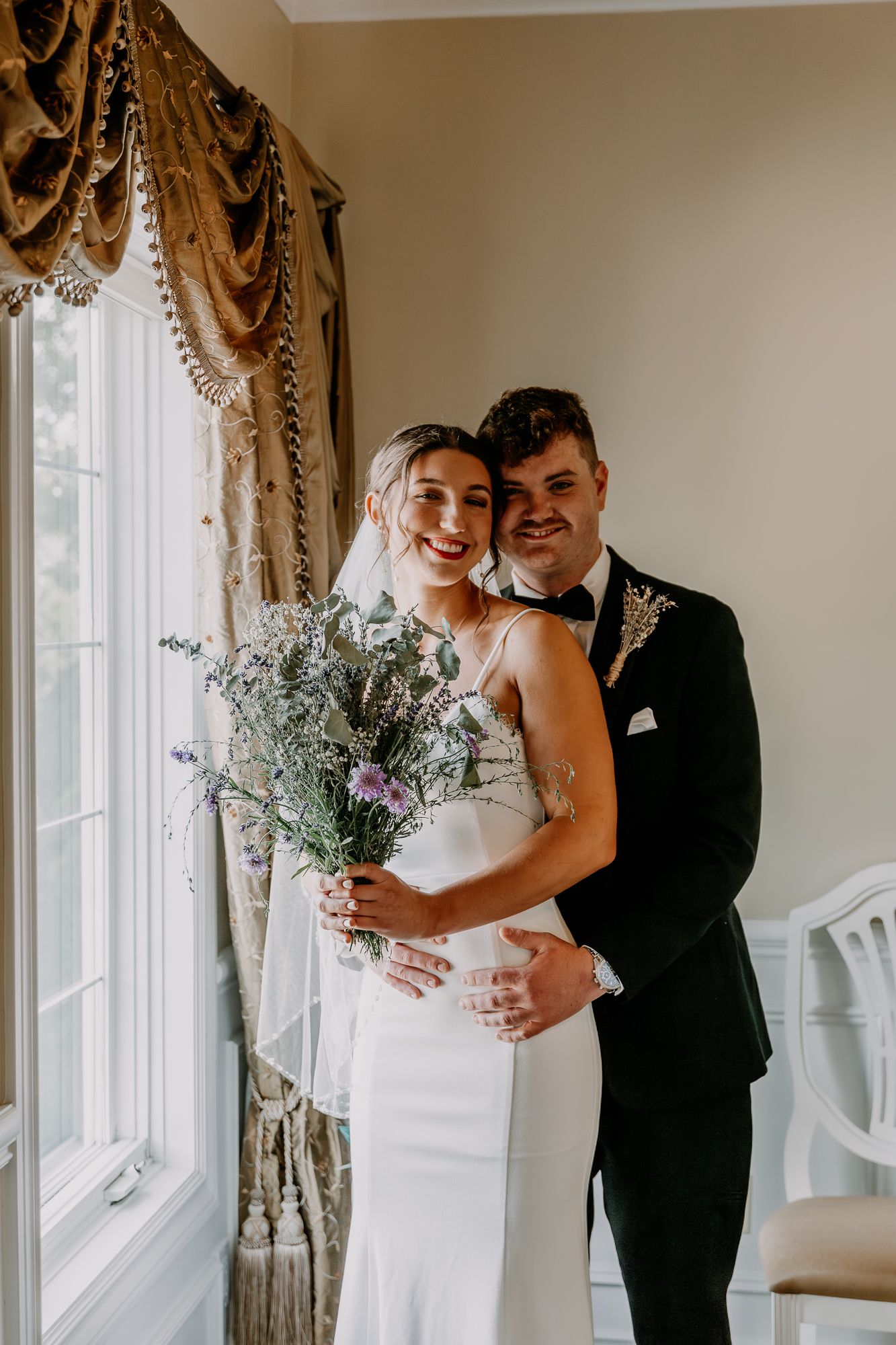 Newlyweds pose together by window with ornate curtains holding wildflower bouquet.