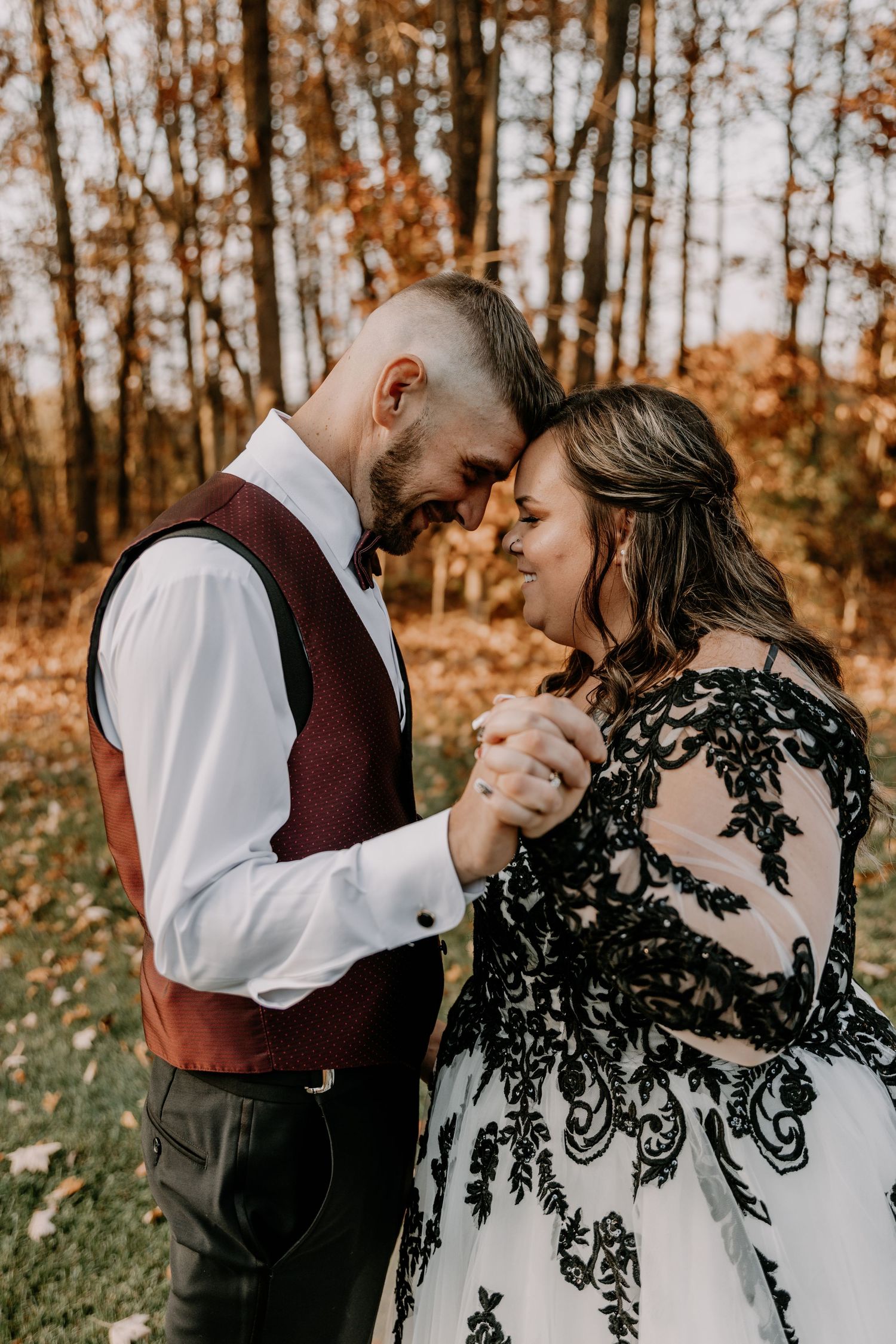 Couple shares romantic moment in fall woodland setting wearing burgundy and black lace wedding attire.