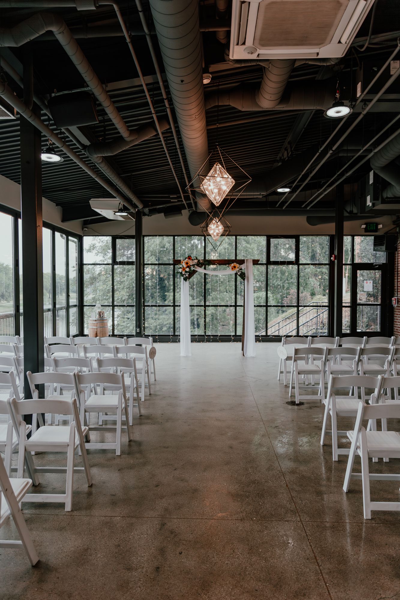 Modern industrial wedding ceremony space with white chairs and large windows.