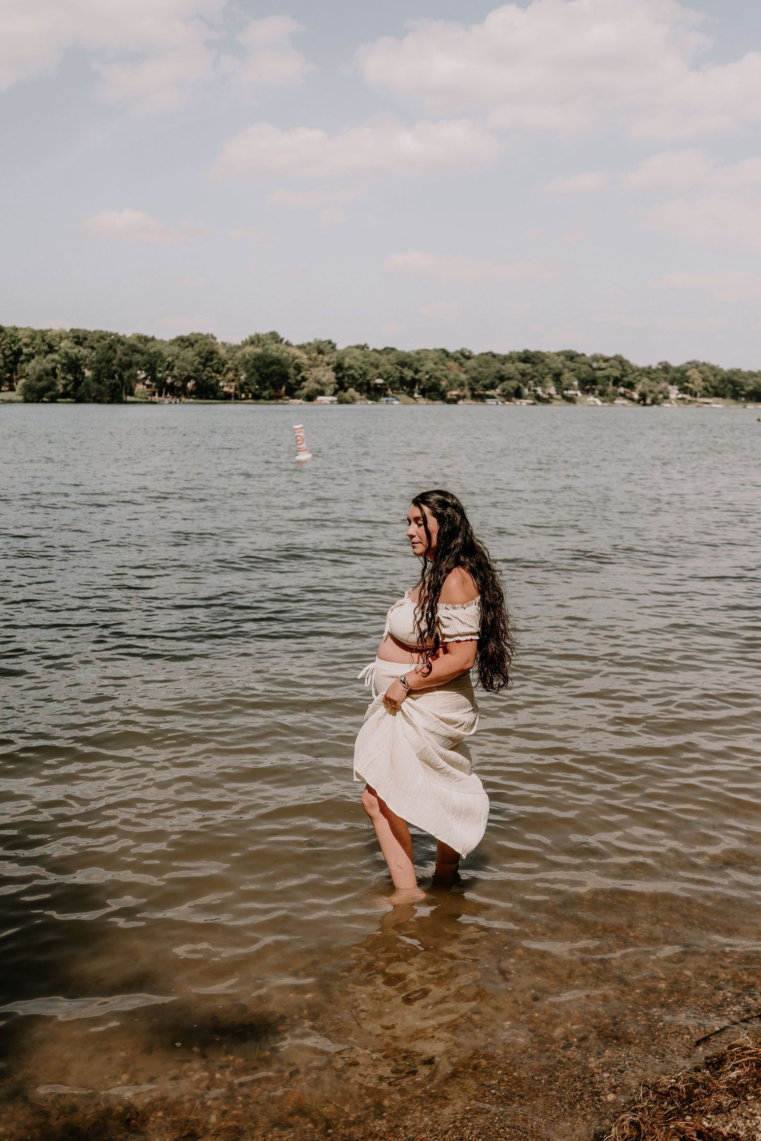 Woman in white dress wading in a calm lake with trees and cloudy sky in the background.