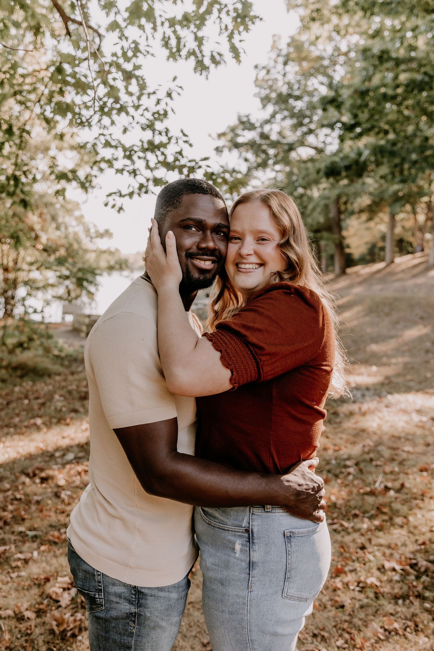 A couple embraces and laughs together while standing in a park with autumn leaves scattered on the ground.