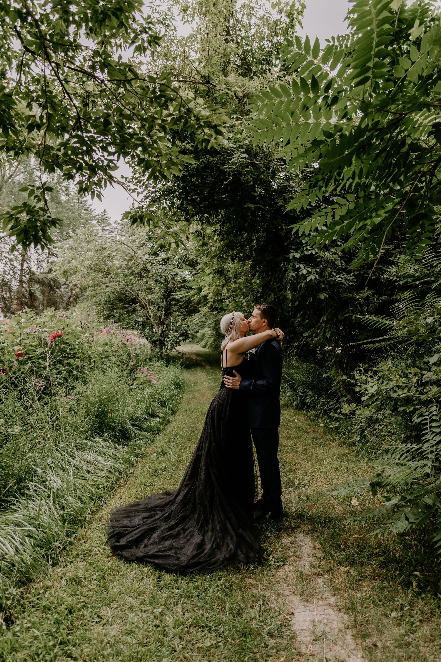 Couple embracing on a garden path, woman in elegant black gown with train, surrounded by lush greenery.