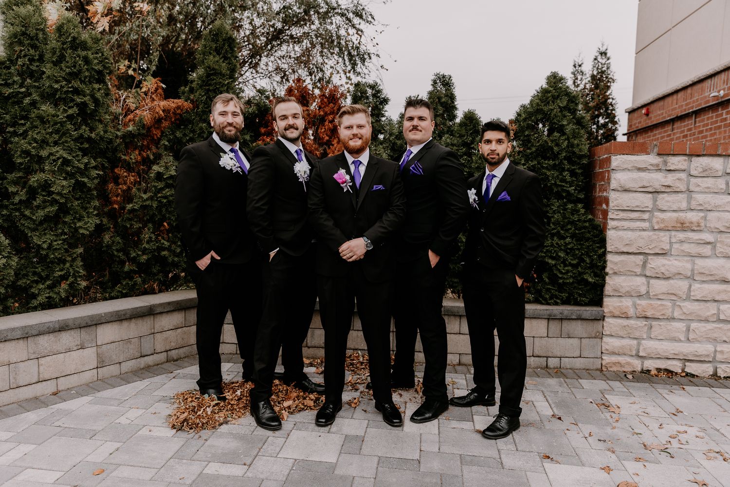 Groomsmen in black suits pose together outdoors against a stone wall and evergreen backdrop.