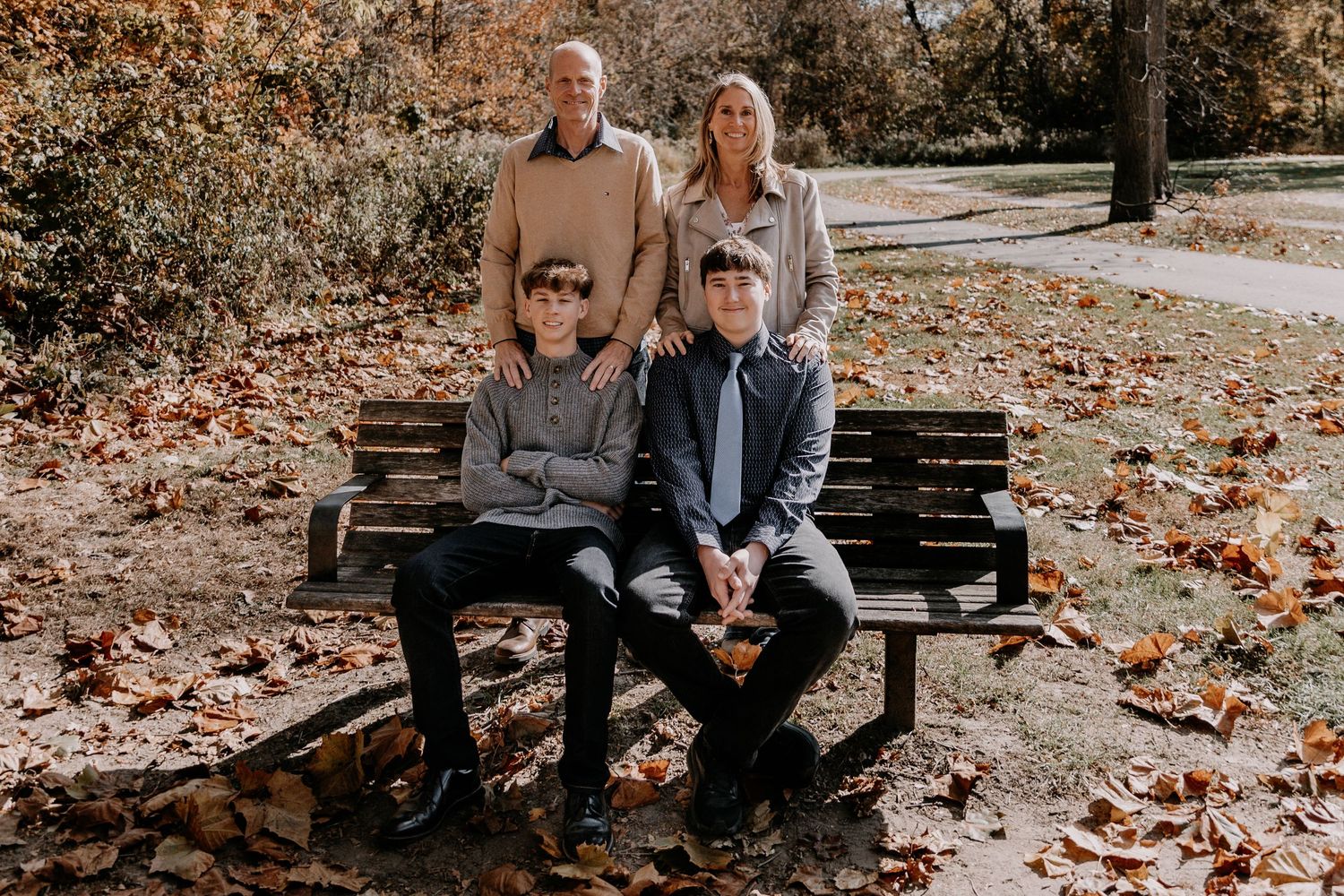 Family of four sitting on wooden park bench surrounded by fallen autumn leaves.