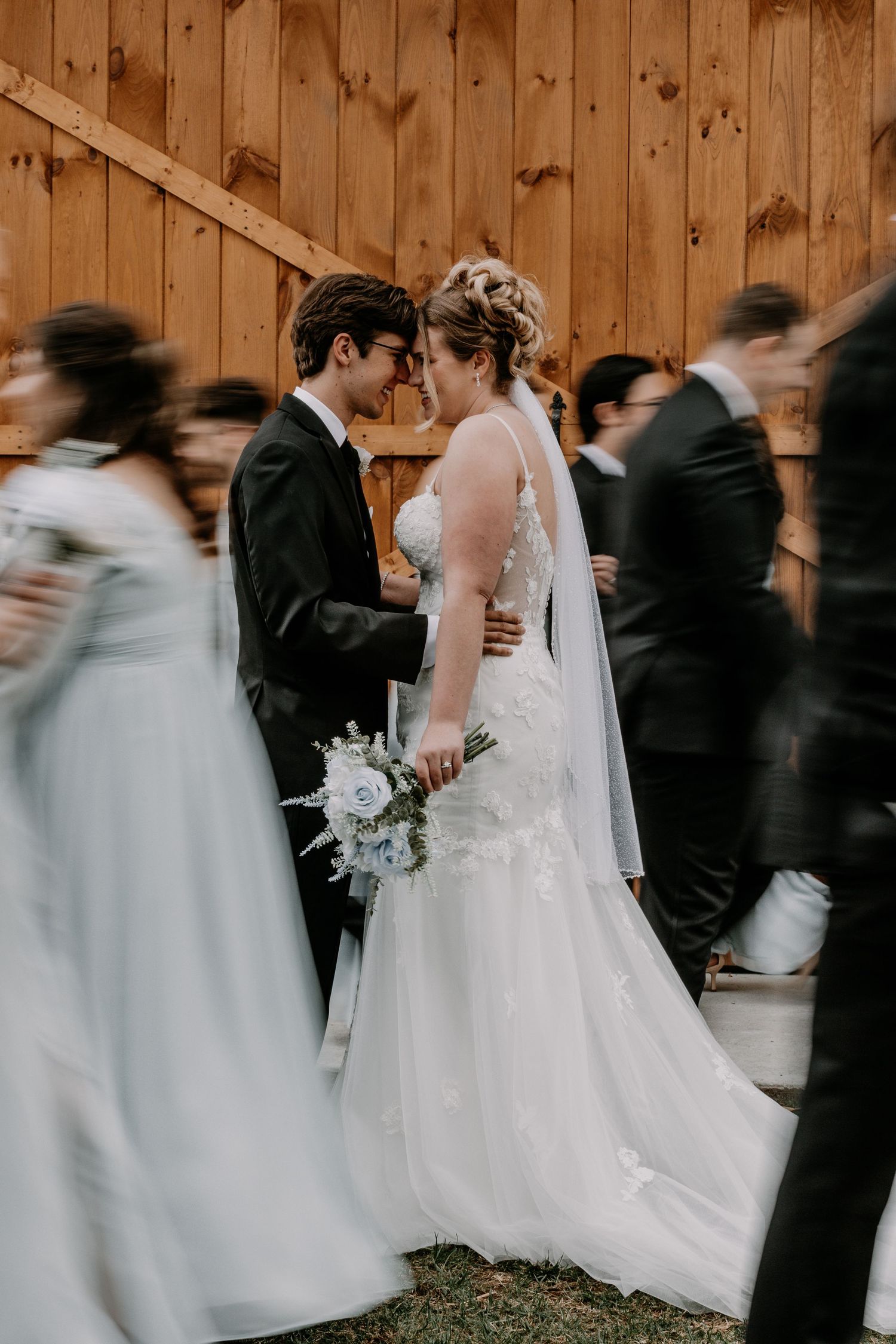 Newlyweds share an intimate moment as wedding guests pass by outside a wooden barn.
