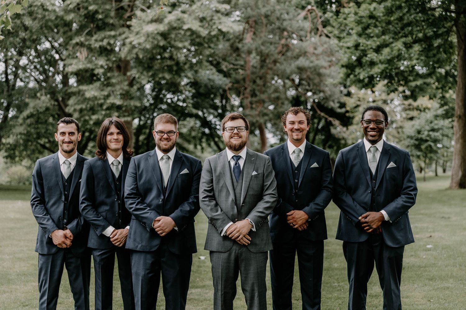 Groomsmen lined up outdoors wearing matching dark suits in garden setting.