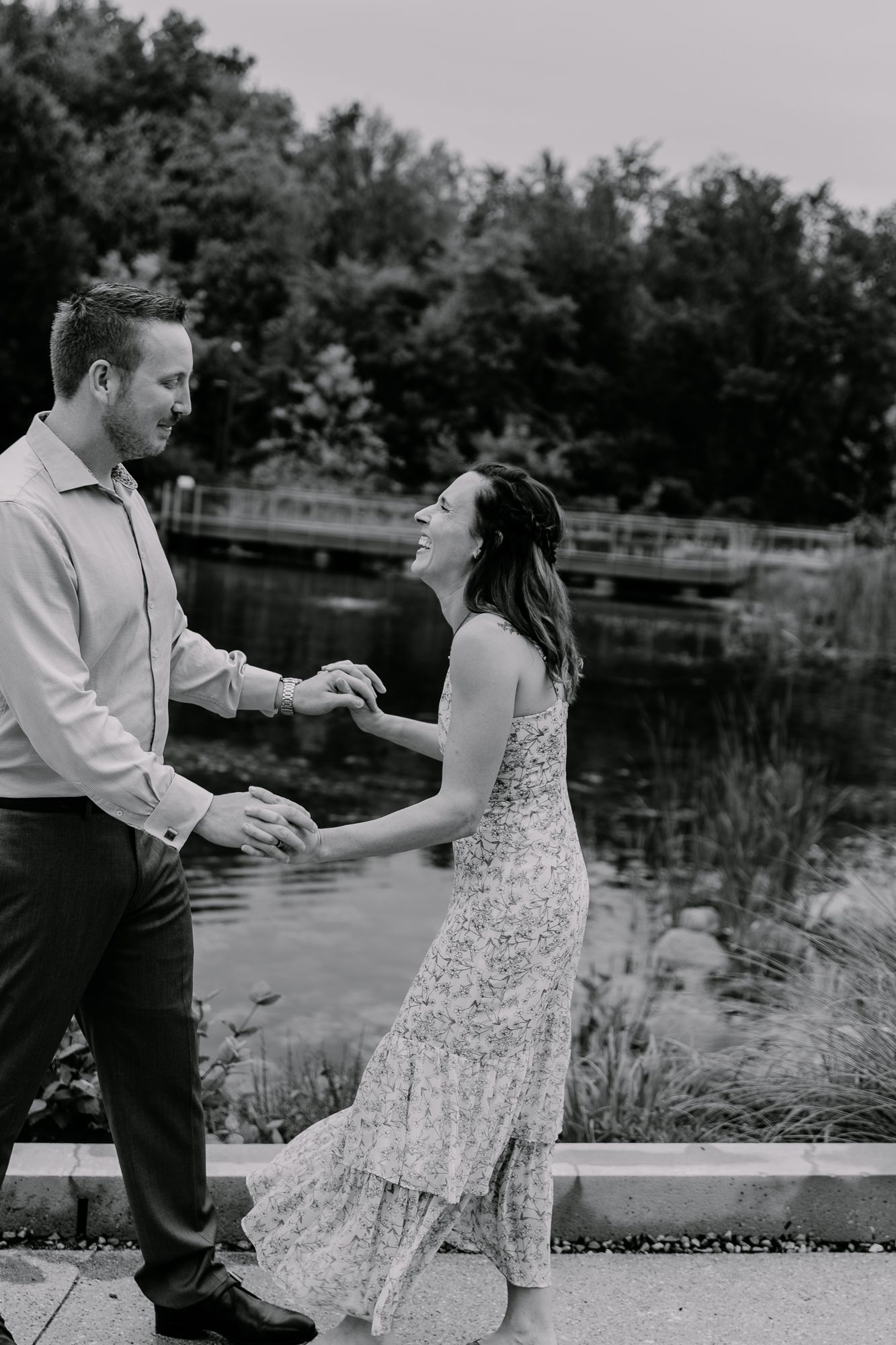 A couple shares a playful moment by a lake, with the woman in a floral dress laughing during their engagement photoshoot.