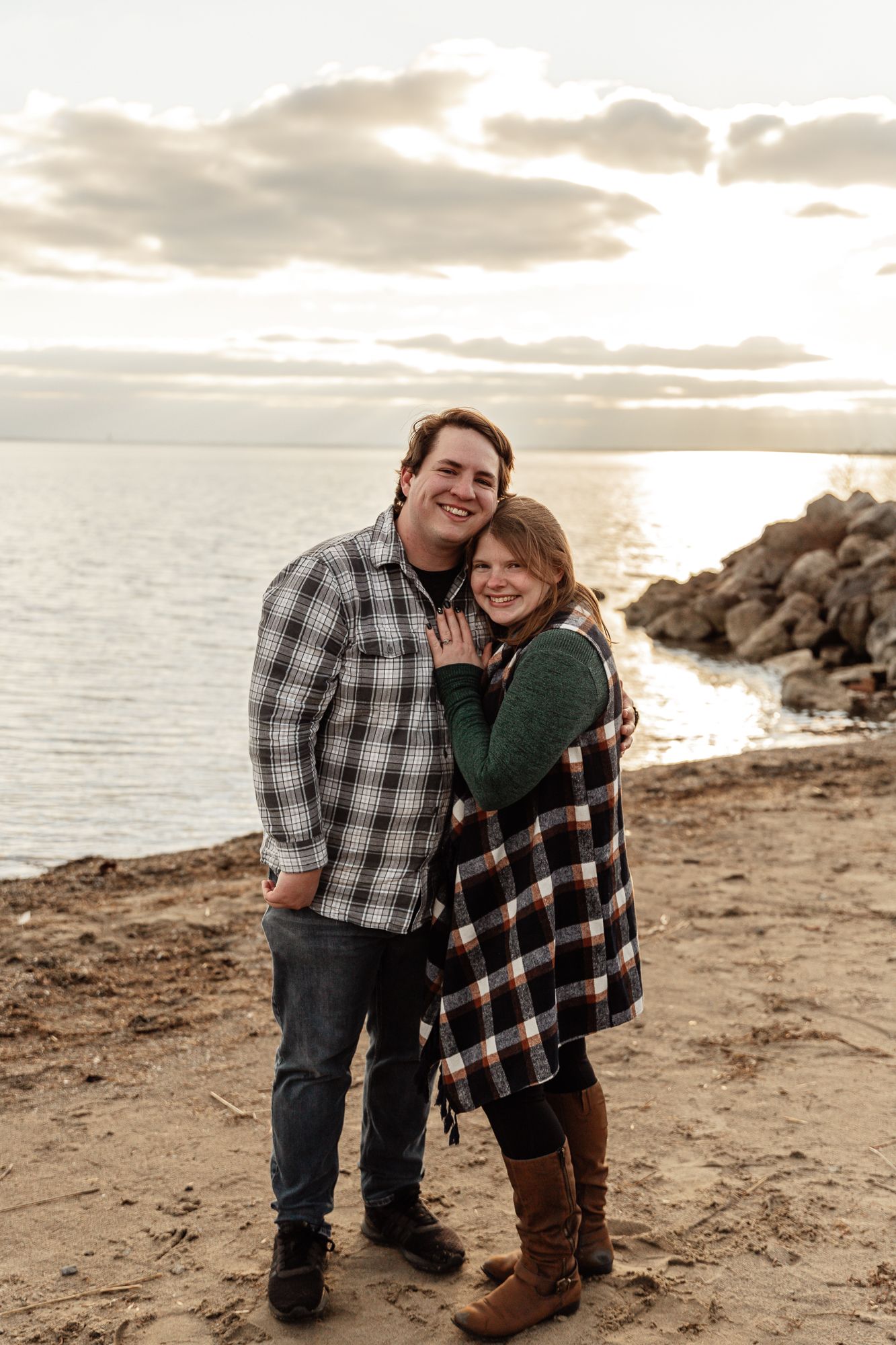 A romantic couple smiling and embracing on a rocky beach at sunset.