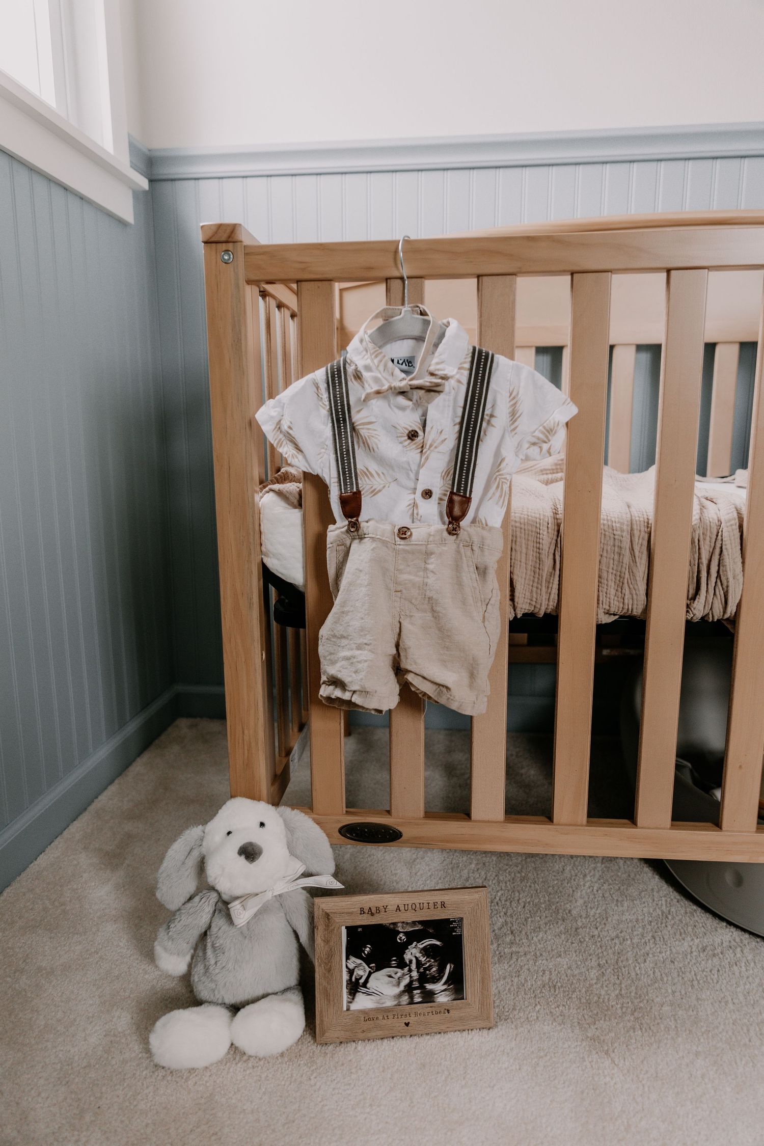 Baby nursery crib with suspender outfit on hanger, ultrasound photo frame, and stuffed bunny toy on carpet floor.