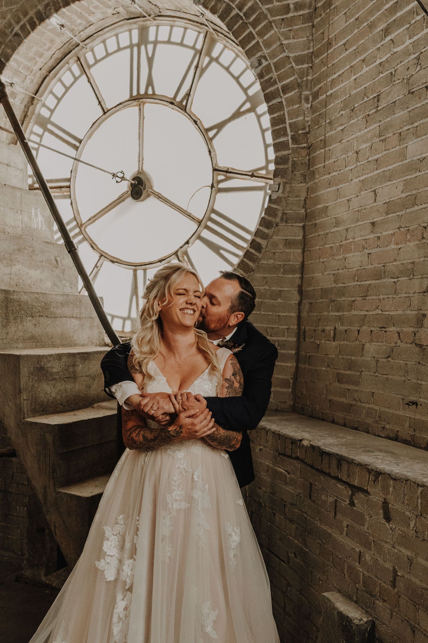 A romantic moment captured in front of a large vintage clock face on exposed brick walls.