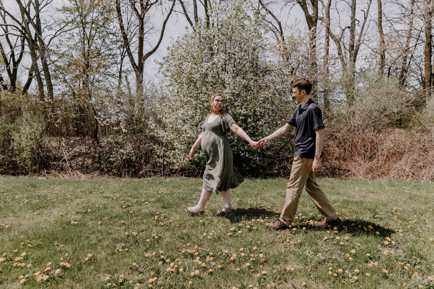 Pregnant woman in green dress holds hands with man in khaki pants, walking through a lush garden with blooming trees.