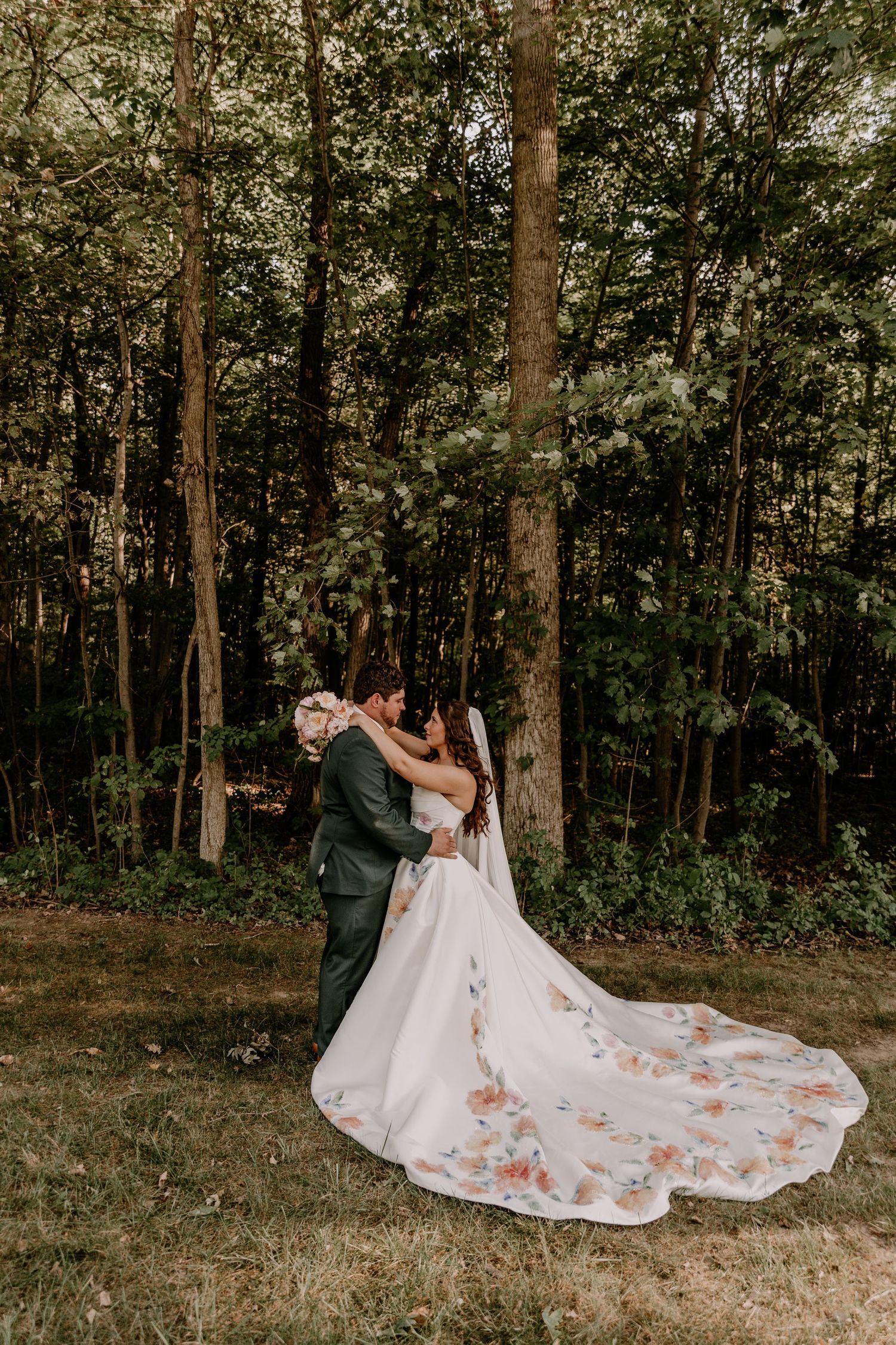 Bride and groom embrace in a forest clearing, her floral-adorned cathedral train spread elegantly on the grass.