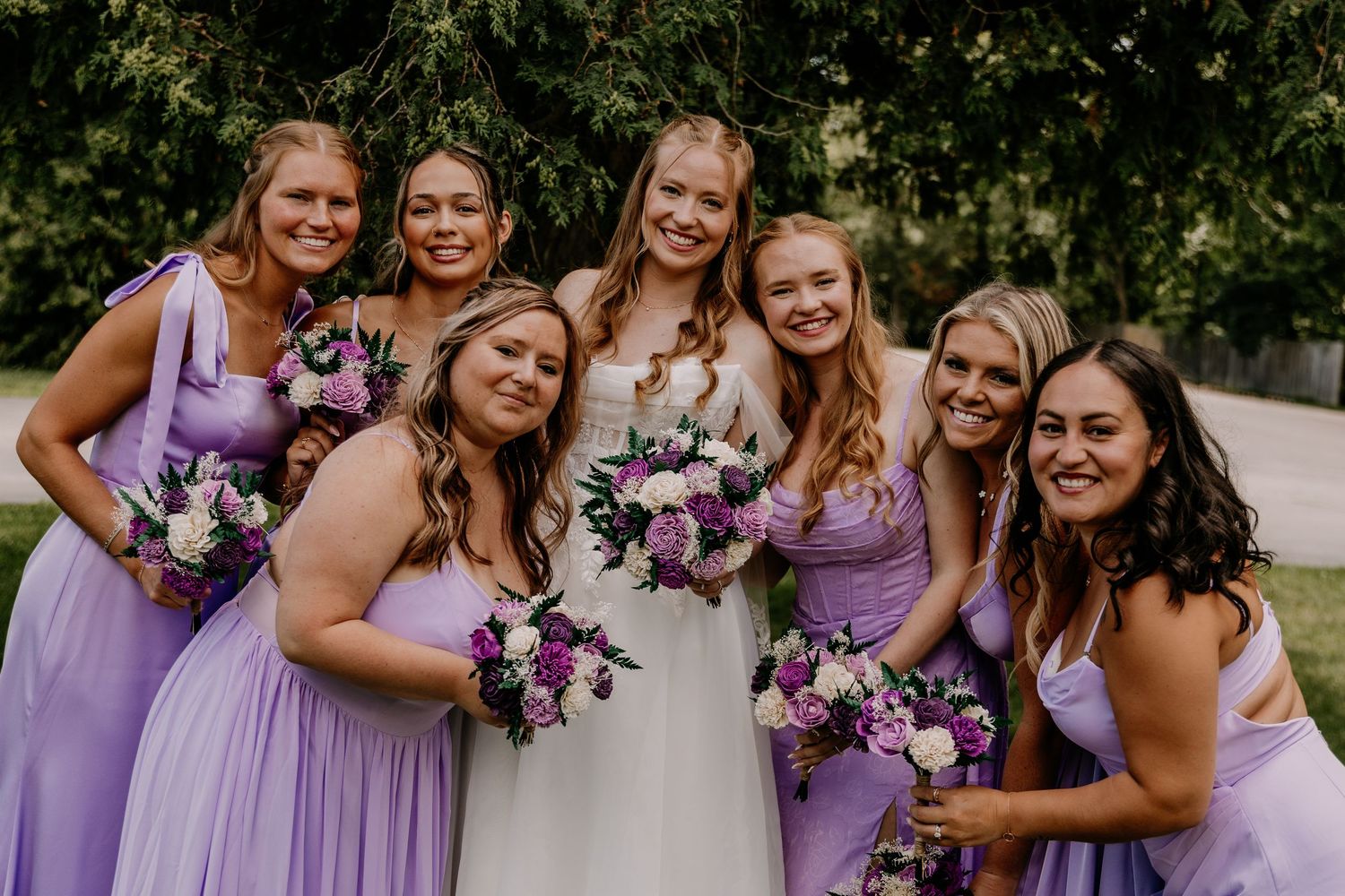 Bride in white gown poses with six bridesmaids in lavender dresses holding purple and white bouquets outdoors.