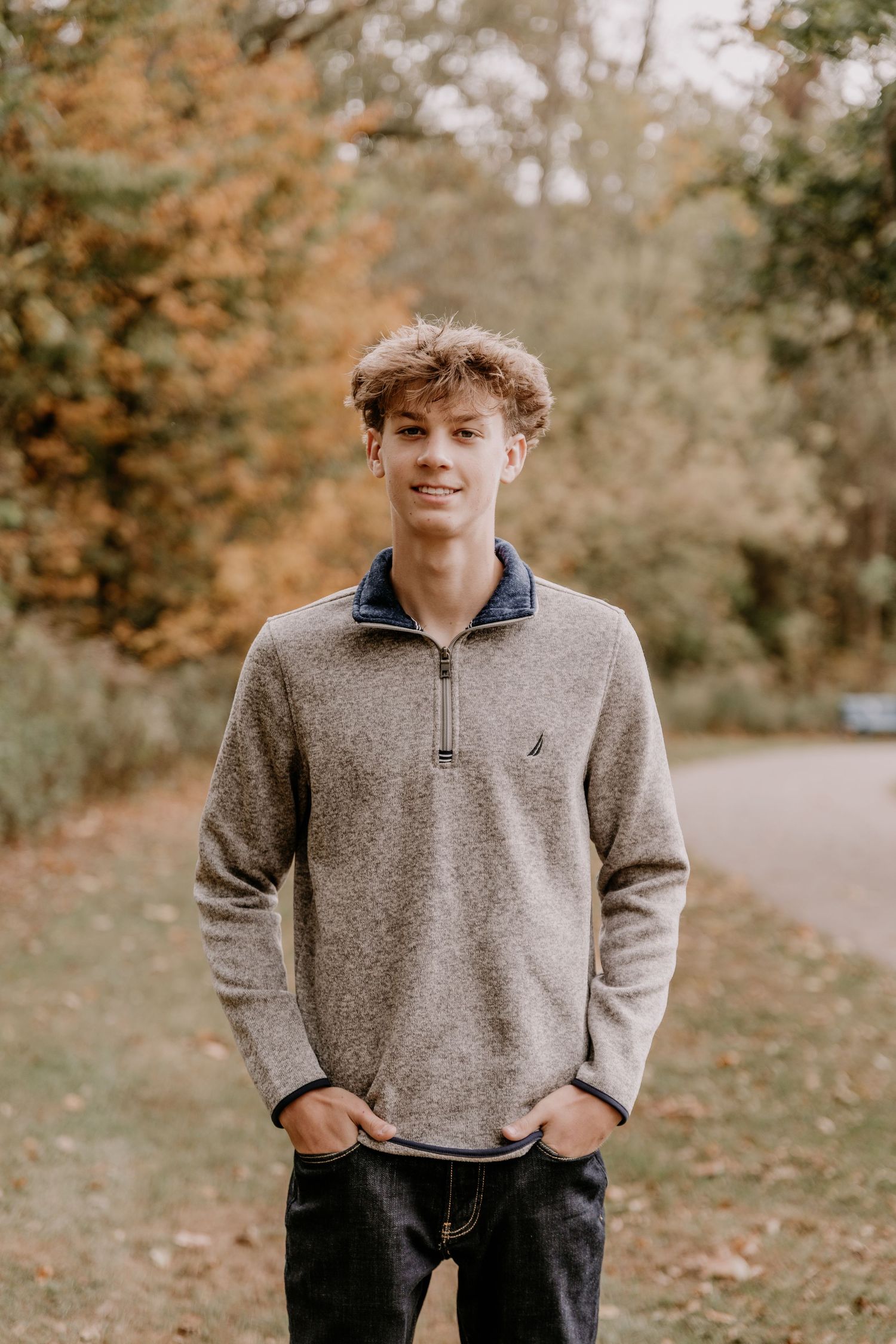 A person in a gray quarter-zip sweater poses outdoors among autumn trees and fallen leaves.