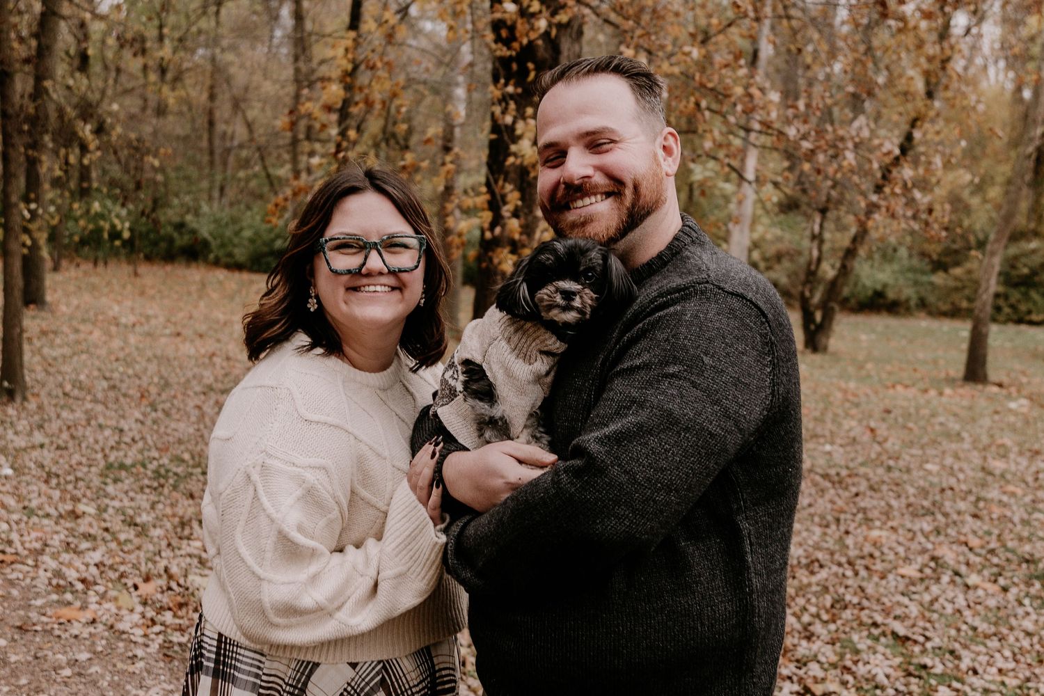 A couple and their small dog pose together in a fall setting with autumn leaves scattered on the ground.